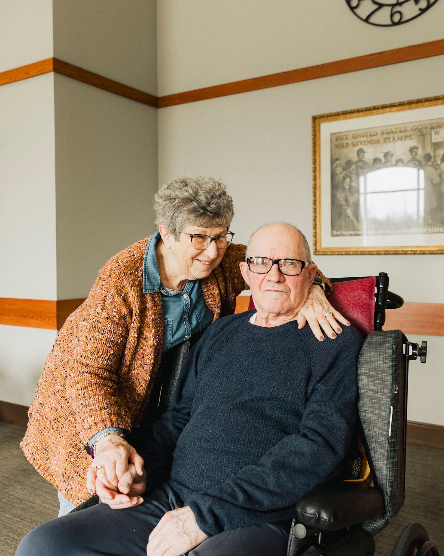 An elderly couple sharing an intimate moment in a room with orange trim and framed artwork.