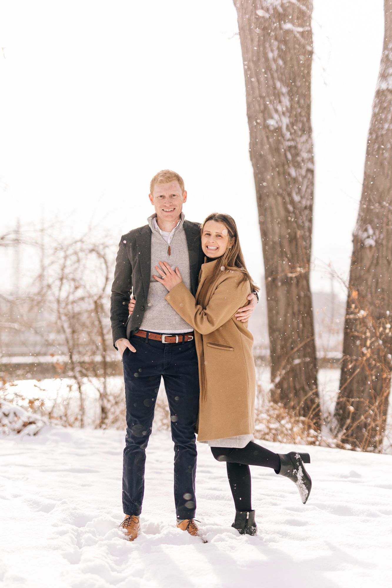 A romantic engagement photo in a snowy park with a couple showing off their ring.