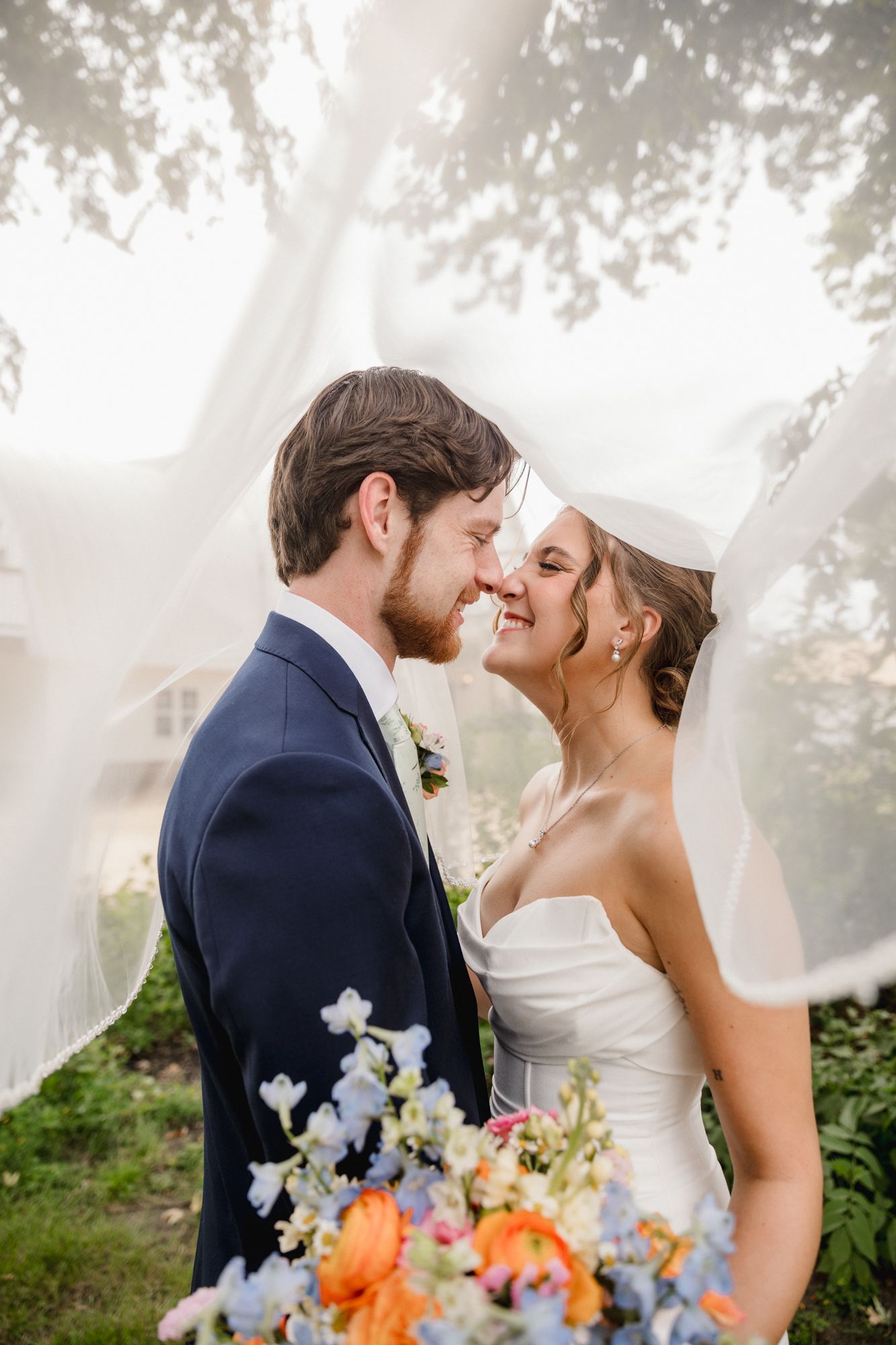 A bride and groom share a romantic nose-to-nose moment under a flowing veil outdoors on their wedding day.
