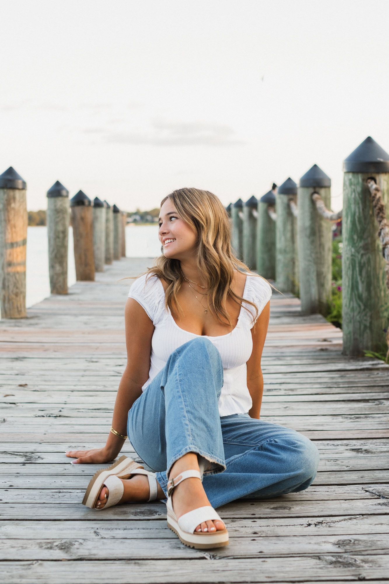 Young woman in white top and jeans sitting on a wooden dock, smiling and looking away, with marina pillars behind her.