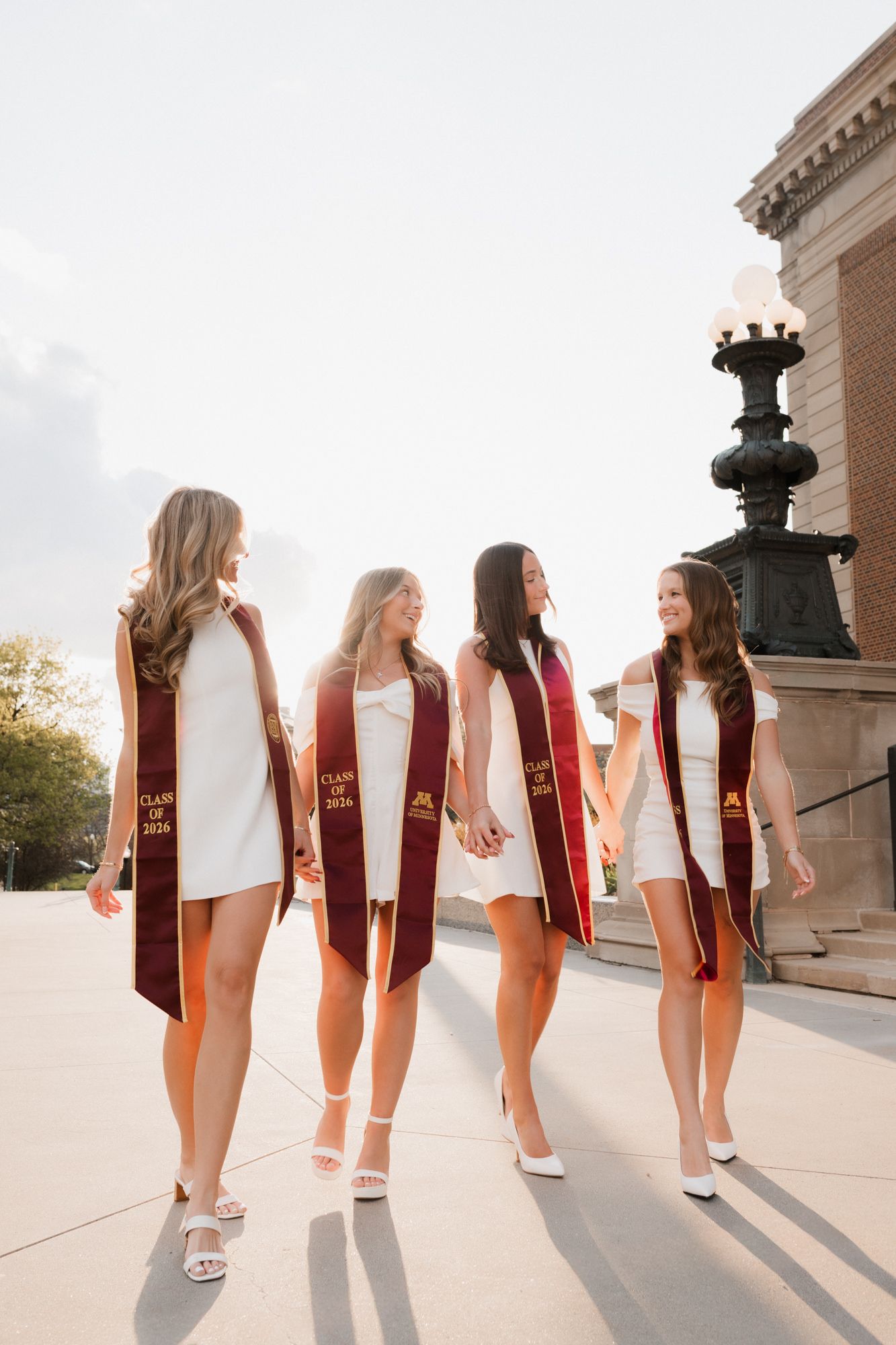 Four University of Minnesota Class of 2024 graduates in white dresses and maroon stoles walking together outdoors.