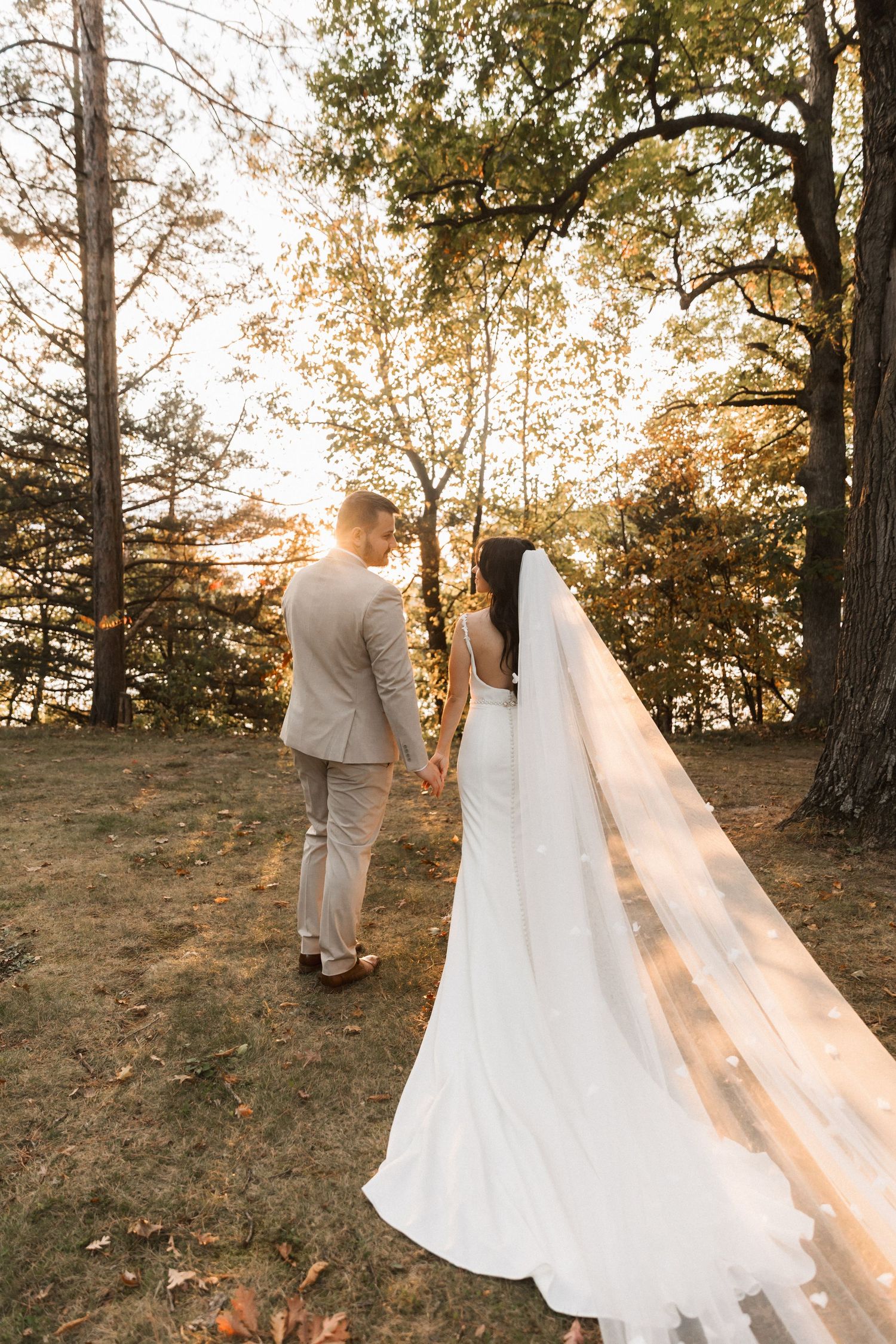 A couple in wedding attire stands together among autumn trees during golden hour sunset.