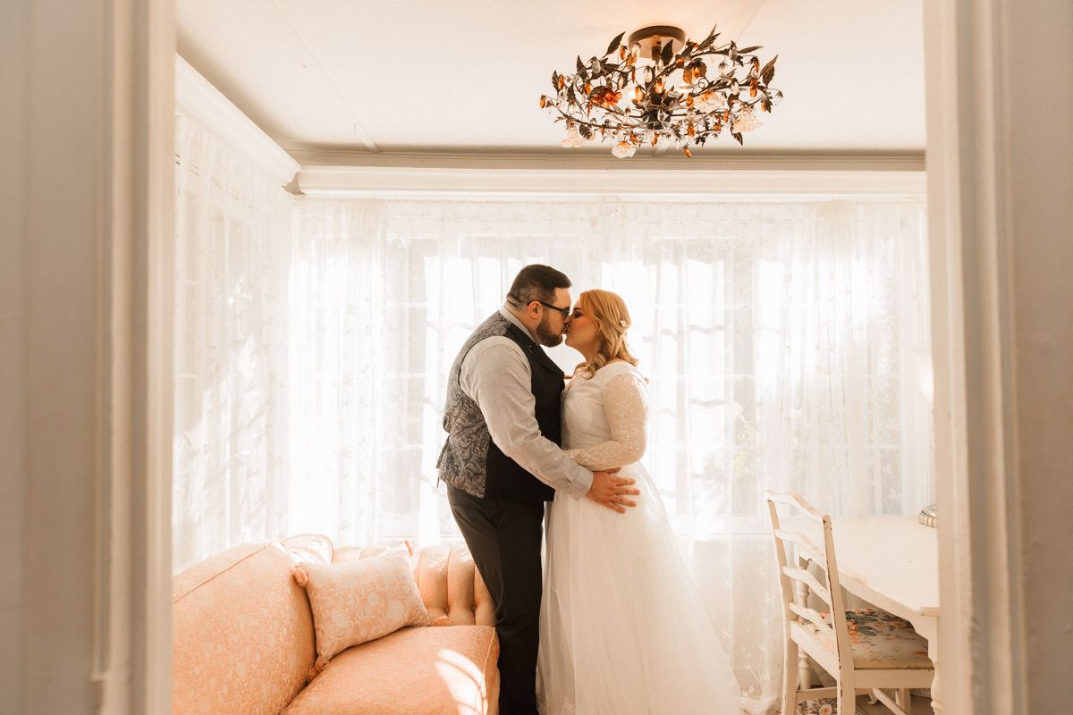 A couple in wedding attire shares an intimate moment in an elegant room with white curtains and chandelier.