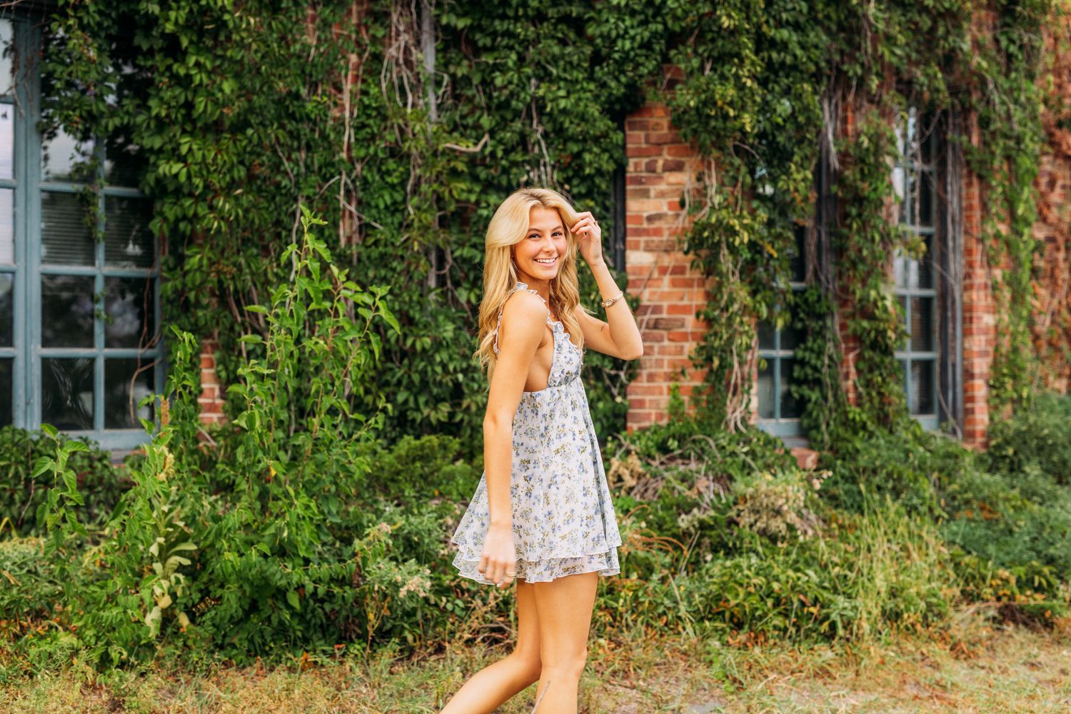 Woman in floral dress strikes playful poses against ivy-covered brick building during outdoor photo shoot.