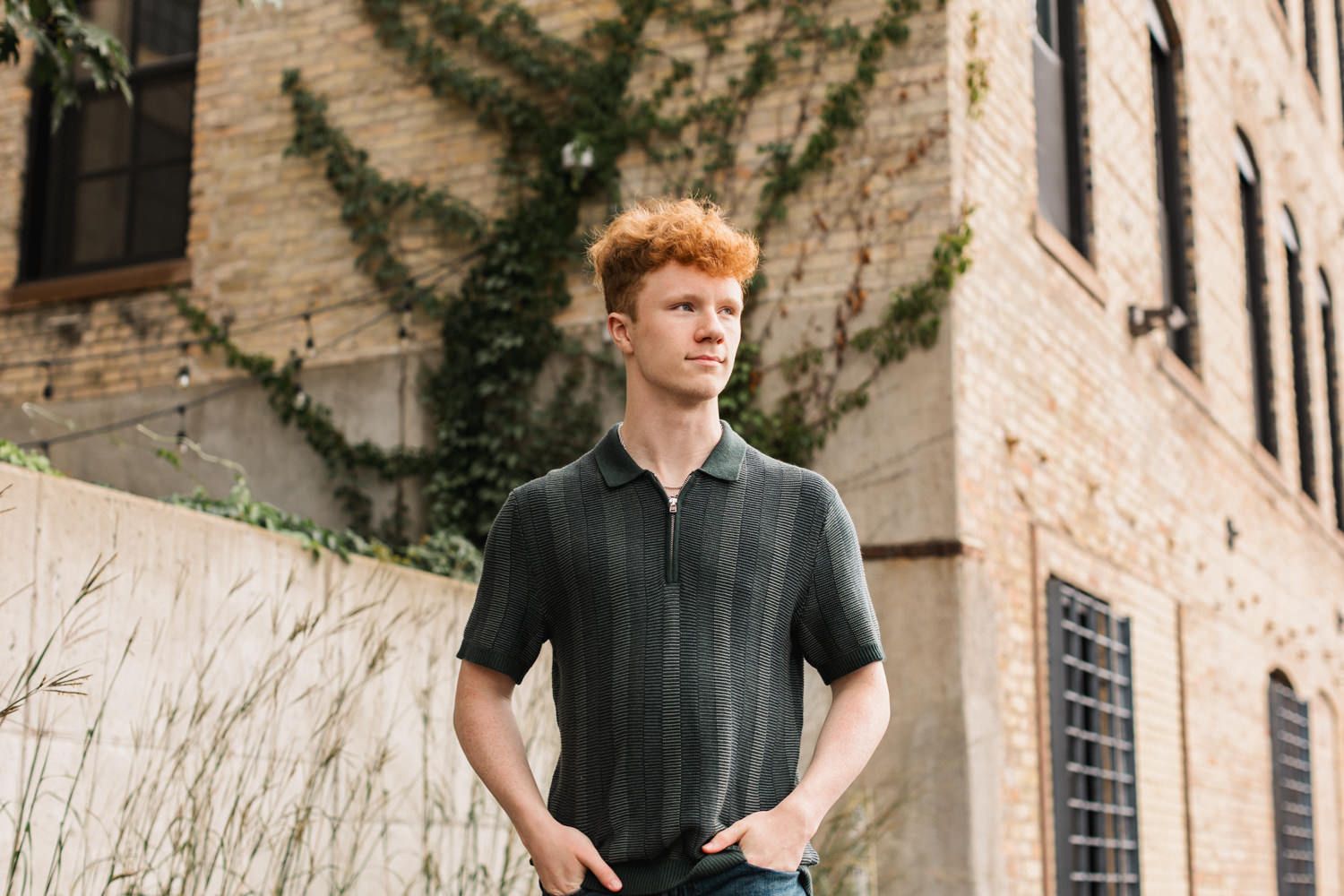 A young person in a dark shirt stands against a brick wall with climbing vines in an outdoor setting.