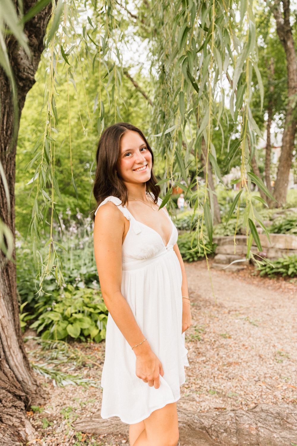 A sunny outdoor portrait under willow trees with someone wearing a flowing white summer dress on a garden path.