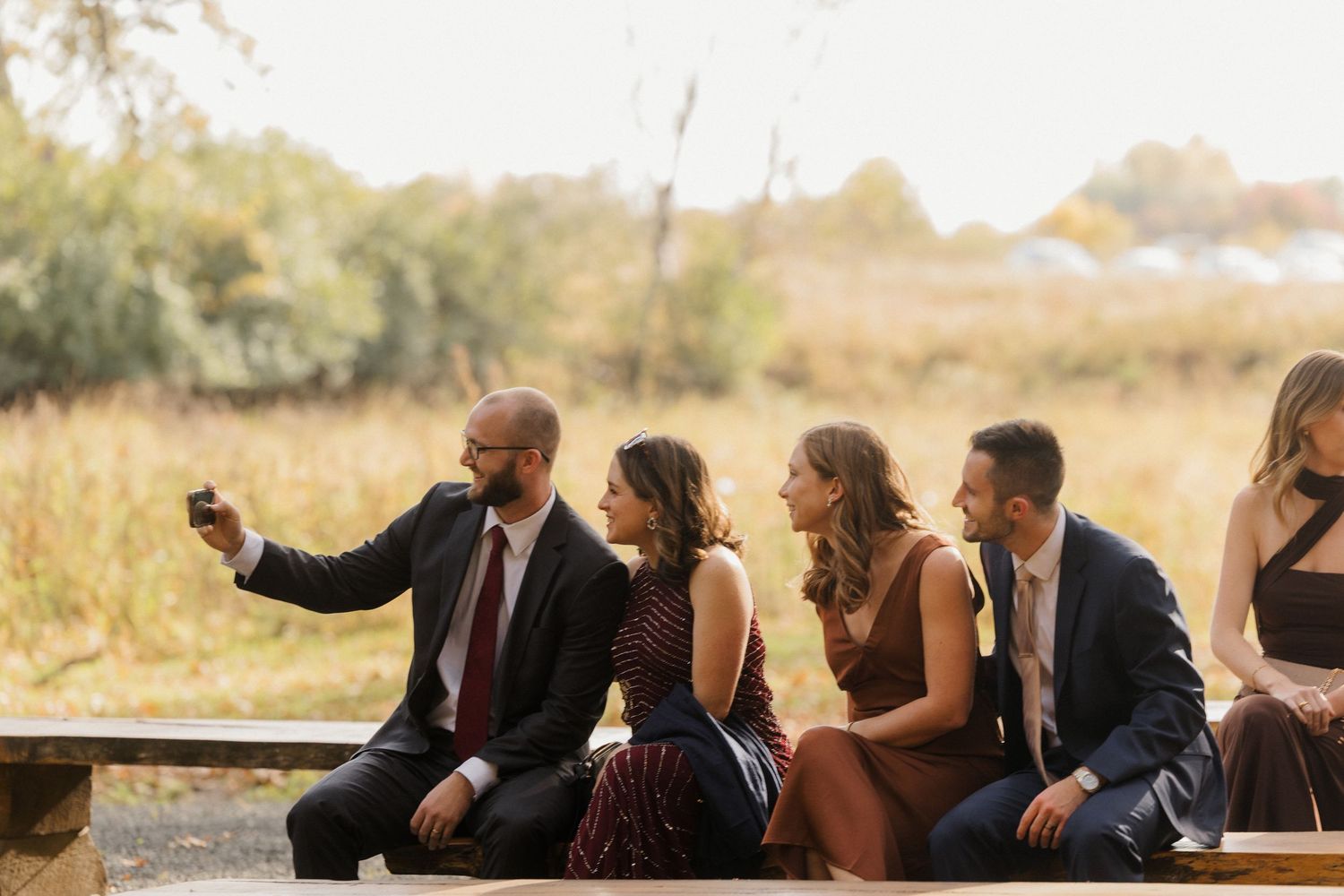 Wedding guests in formal attire sit on a wooden bench beside a golden field while one man takes a selfie with his phone.