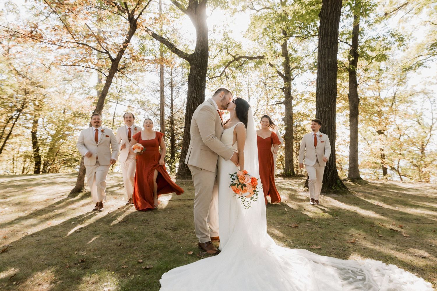 Bride and groom share a kiss outdoors as wedding party in rust bridesmaid dresses and tan suits walk among autumn trees.