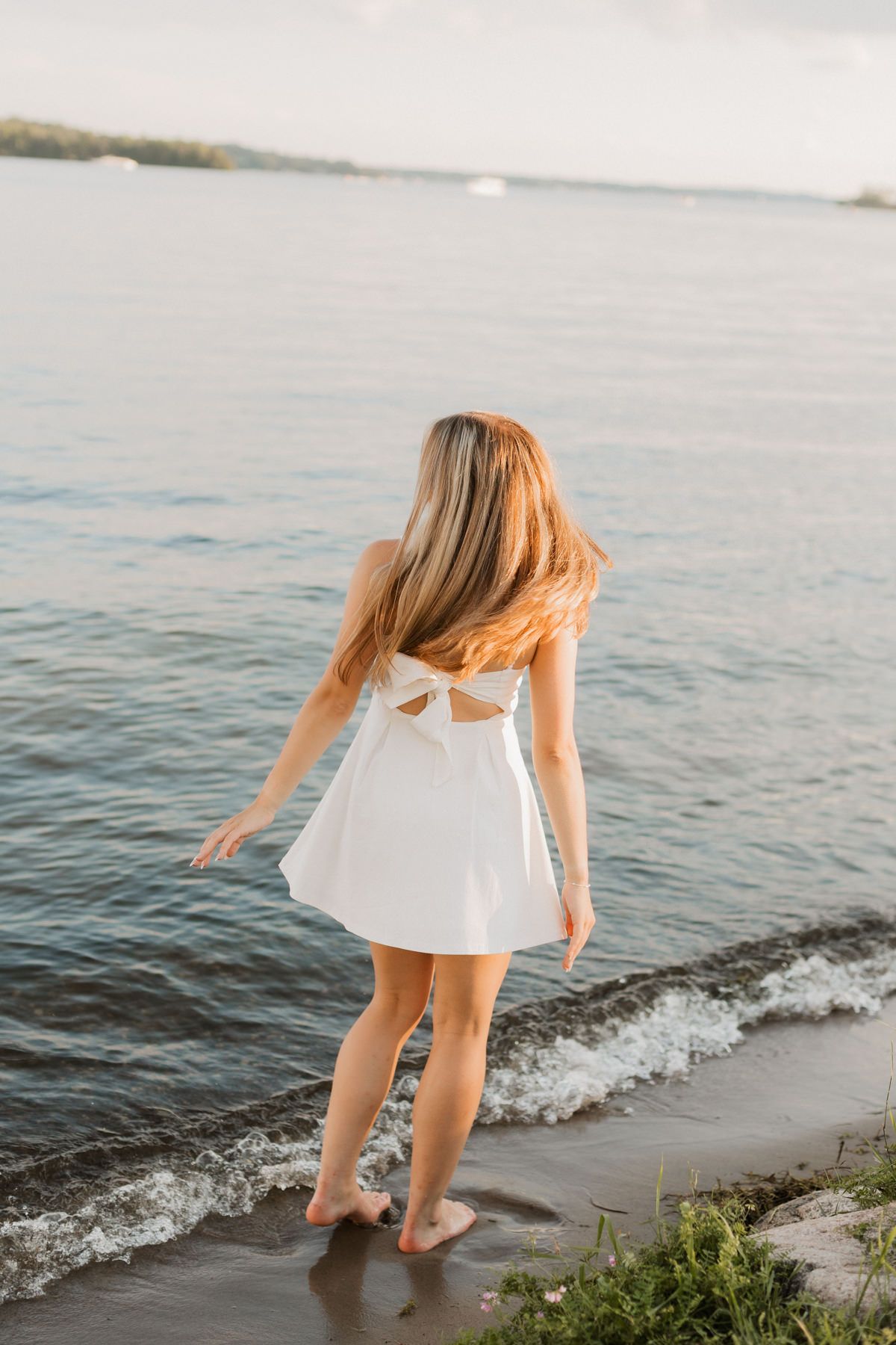 Young woman in white backless dress standing barefoot at water's edge on sandy beach at sunset.