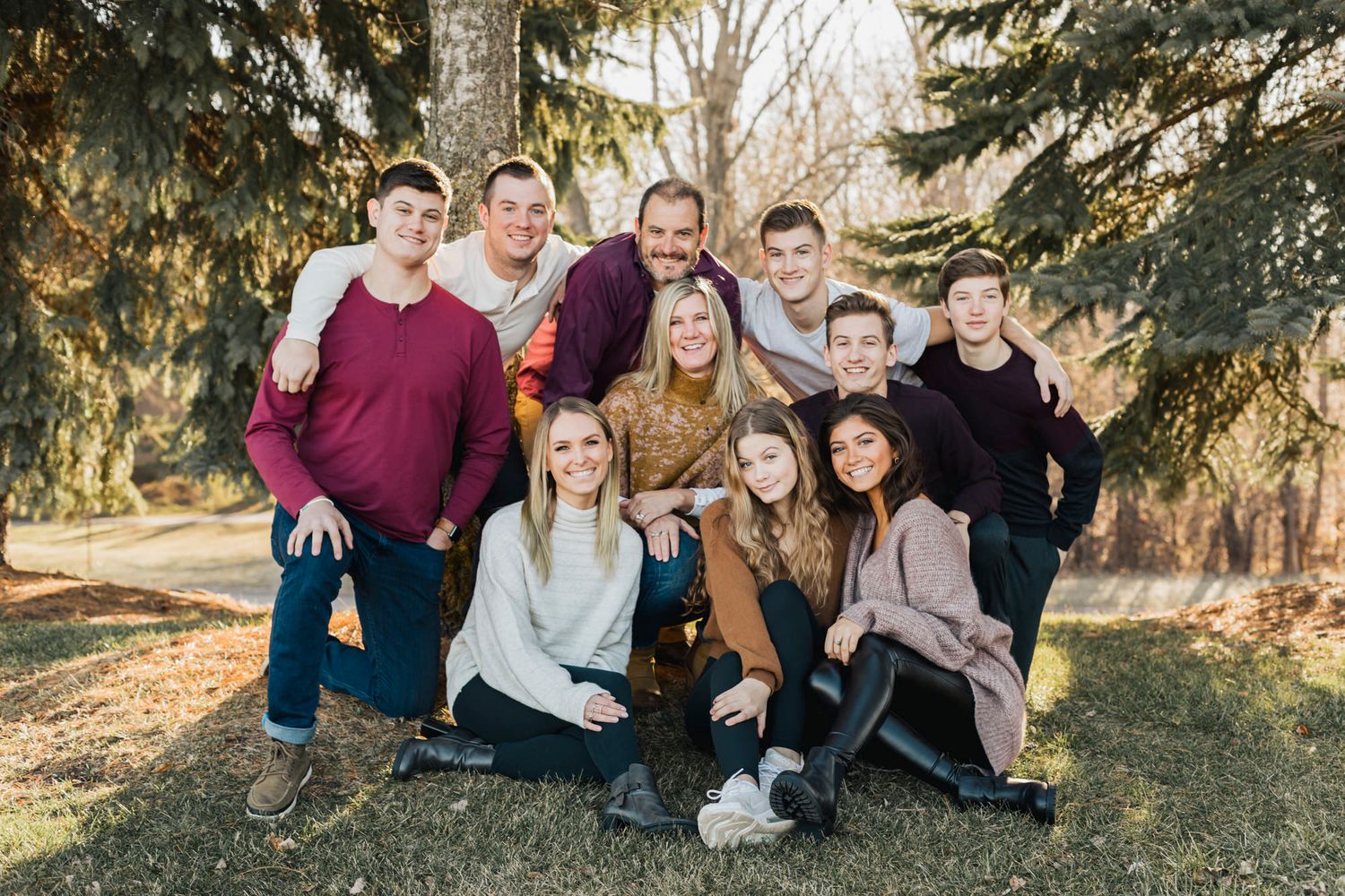 A large group poses together outdoors among evergreen trees in casual attire.