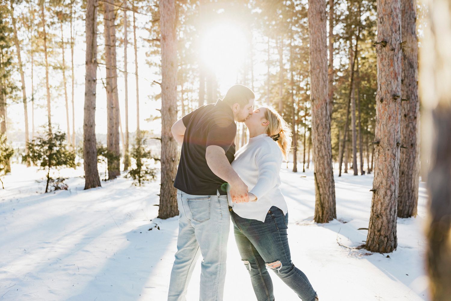 Sun flares through snowy pine trees as a couple shares an intimate moment in winter wonderland.