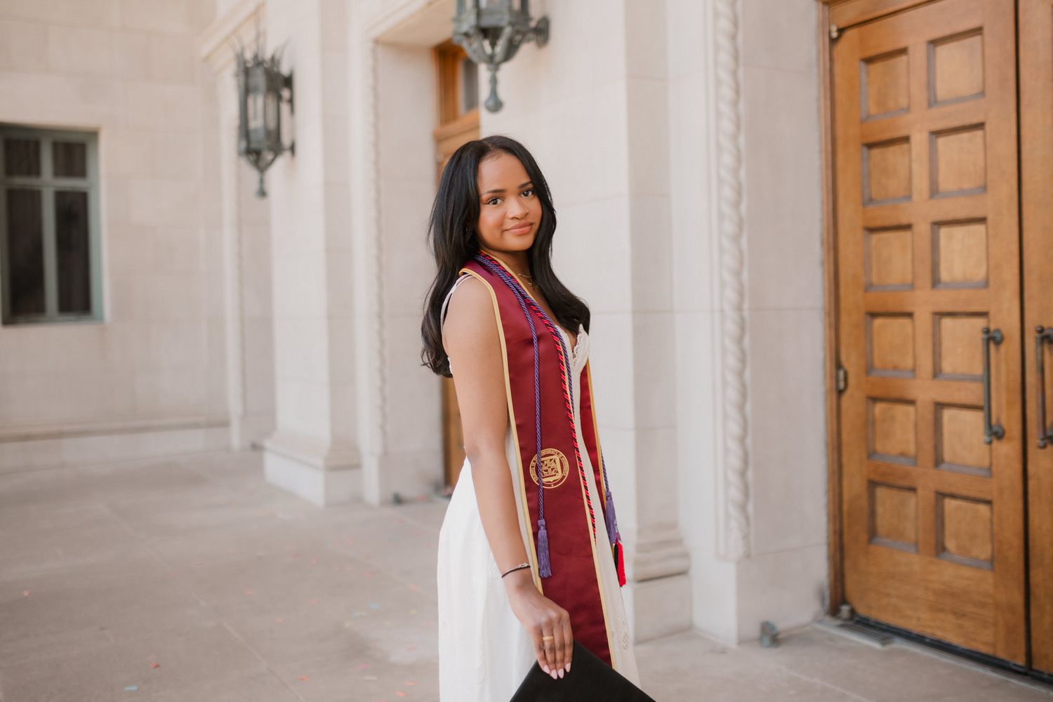 Young woman in white dress and maroon graduation stole smiling outdoors near ornate wooden door.