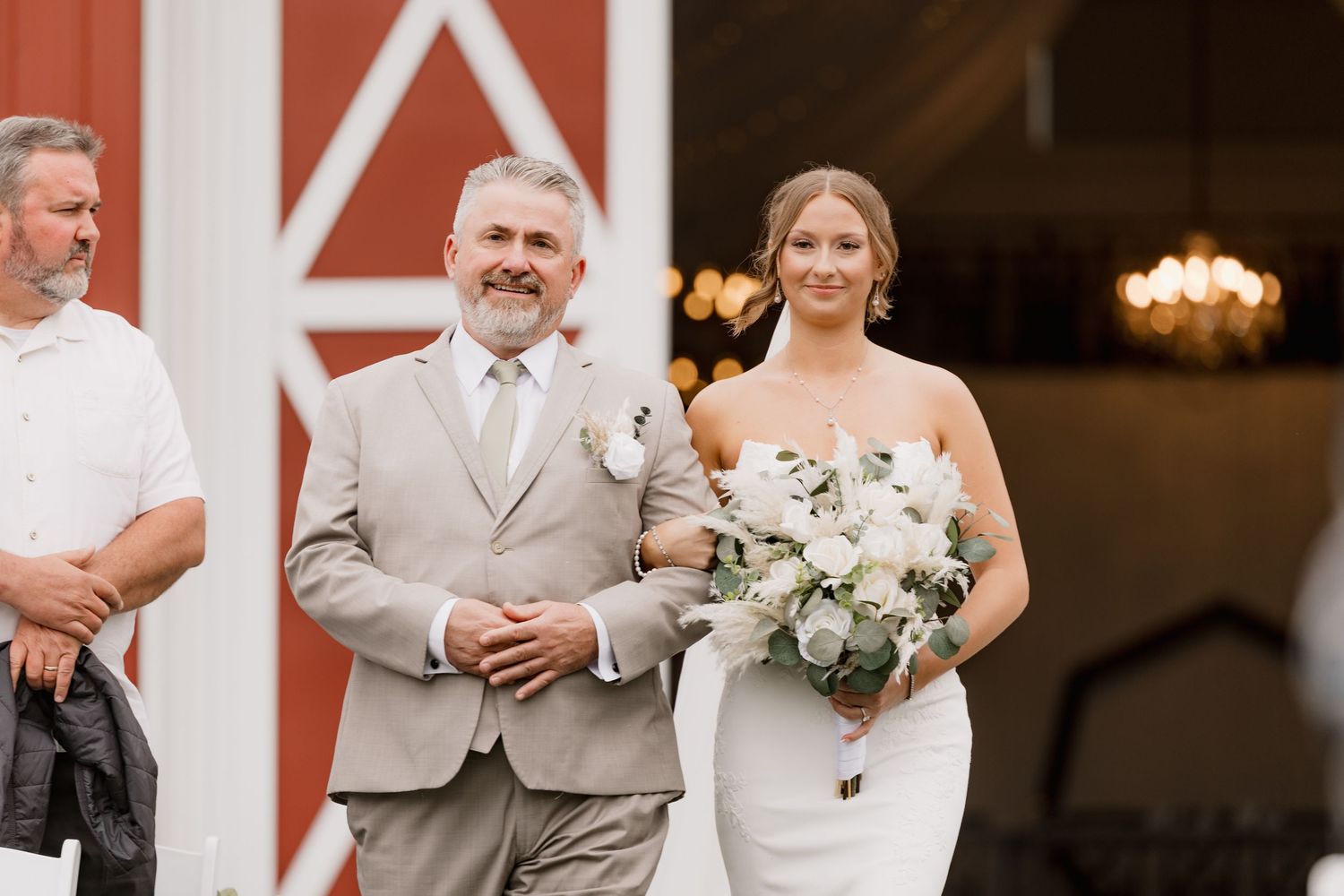 Bride in white gown holding bouquet walks down aisle with escort near red barn wedding venue.
