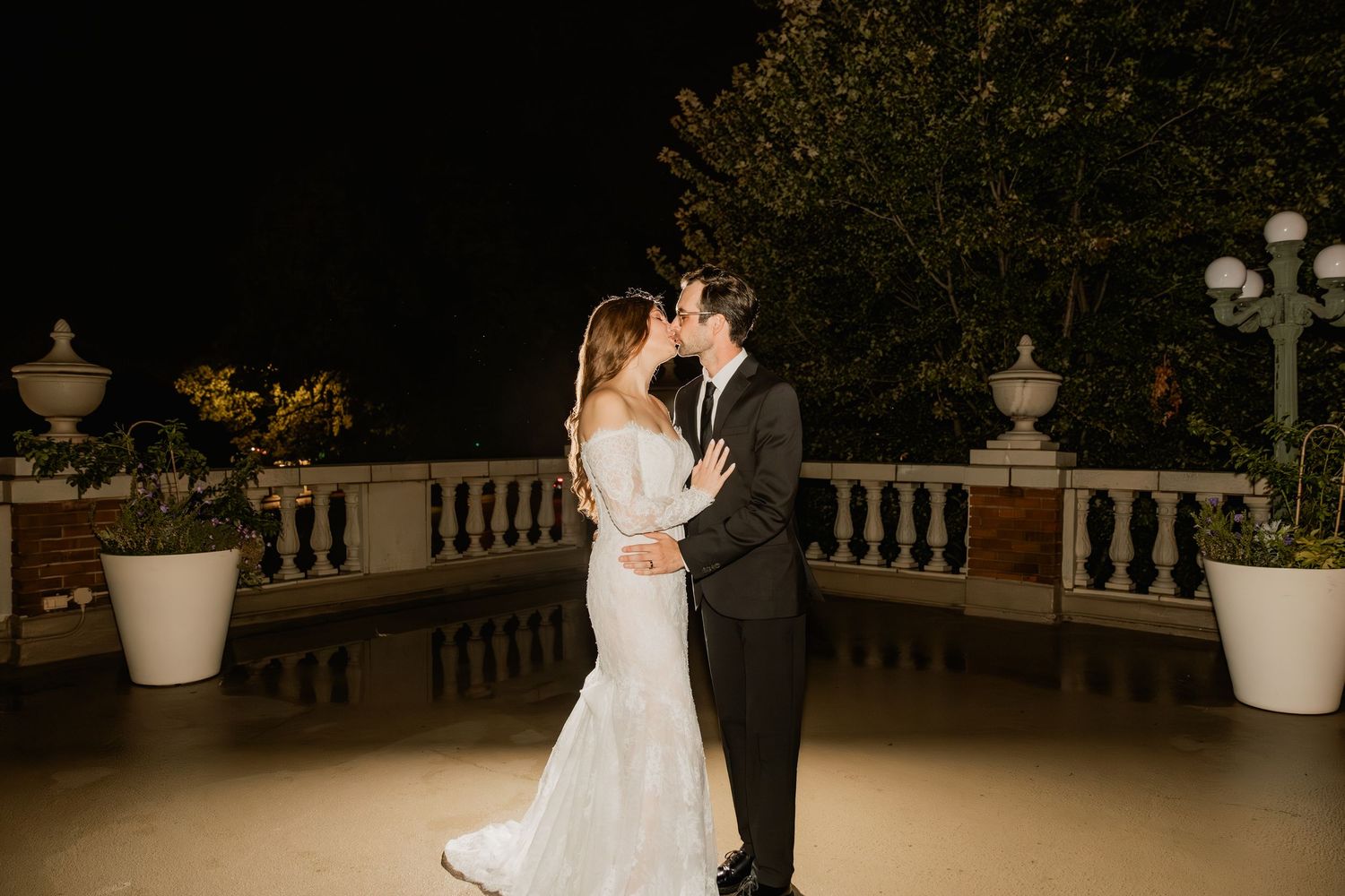 Bride and groom share a romantic kiss on an elegant outdoor terrace at night, surrounded by white planters and lush greenery.