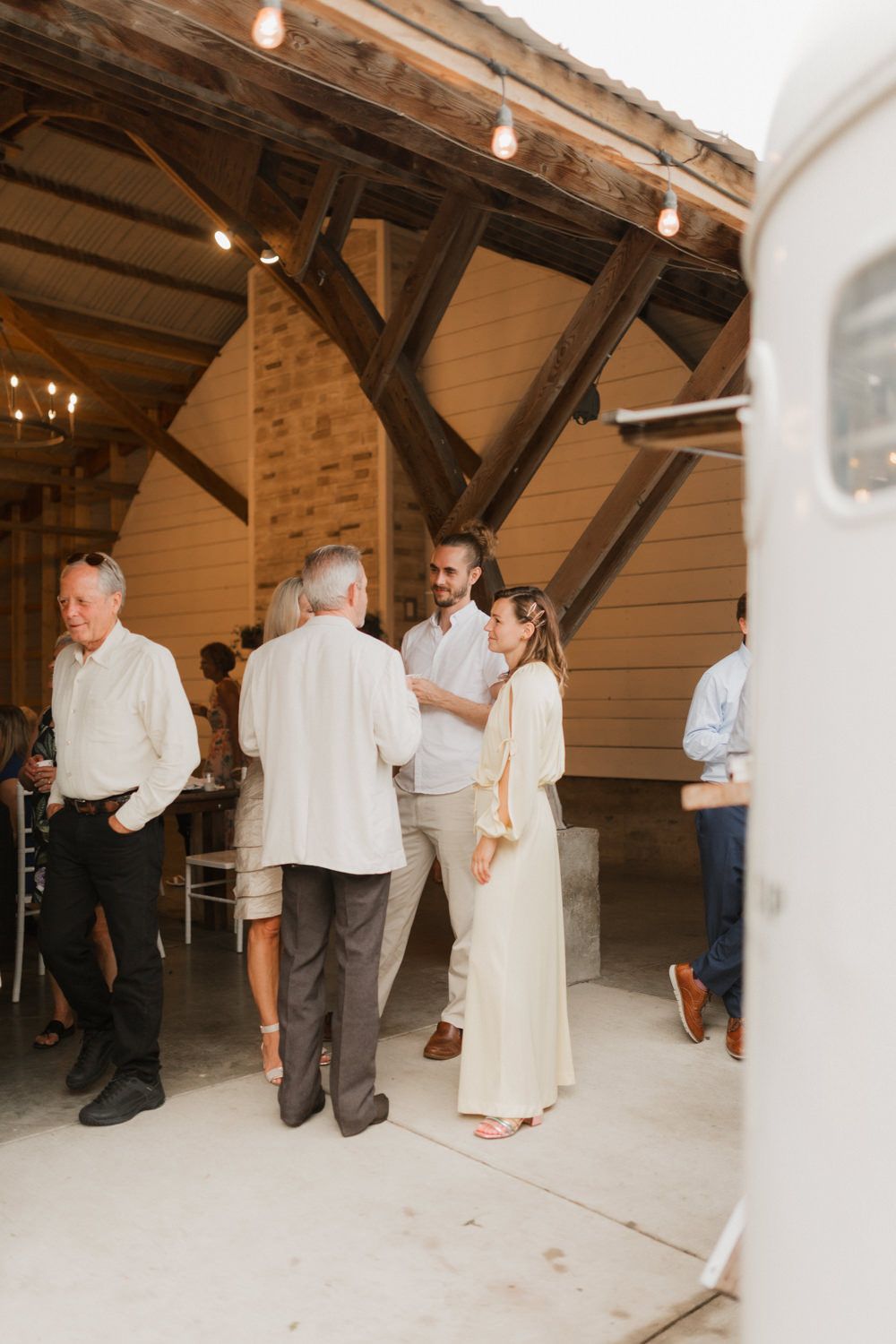 People in white and gray attire gather in a rustic attic space with exposed wooden beams and hanging lights.