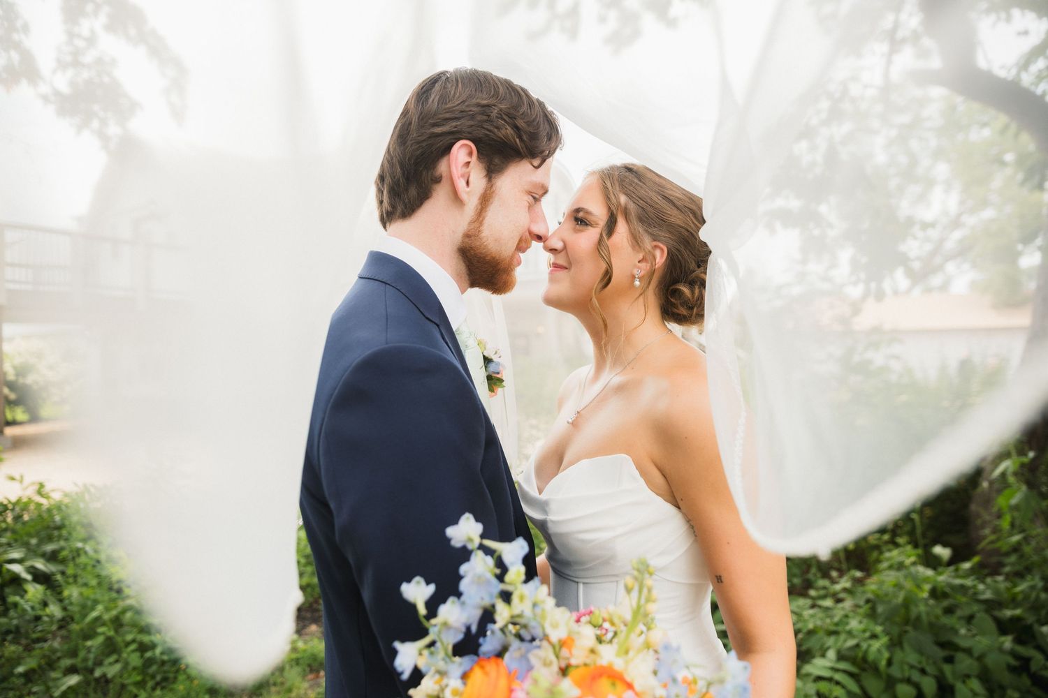 Bride and groom share a romantic moment under a flowing veil, surrounded by lush greenery on their wedding day.