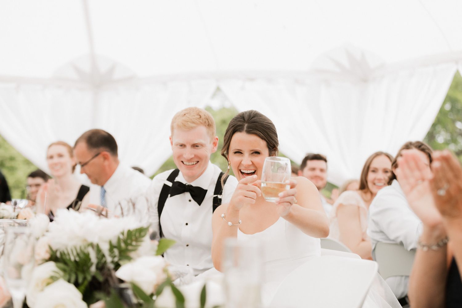 Bride and groom toasting champagne at elegant outdoor wedding reception under white tent with guests.