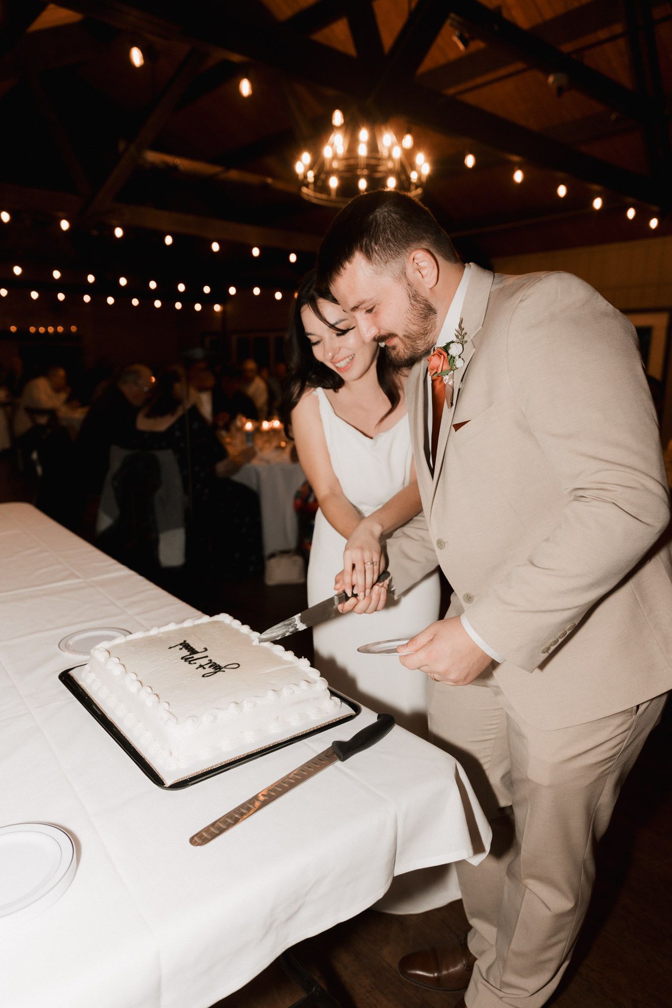 Bride and groom cutting white wedding cake together at rustic reception venue with warm string lights and chandelier.
