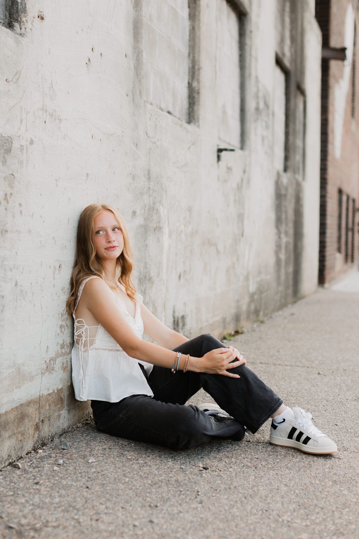 Young woman in white top and black jeans sitting against urban concrete wall wearing Adidas sneakers.