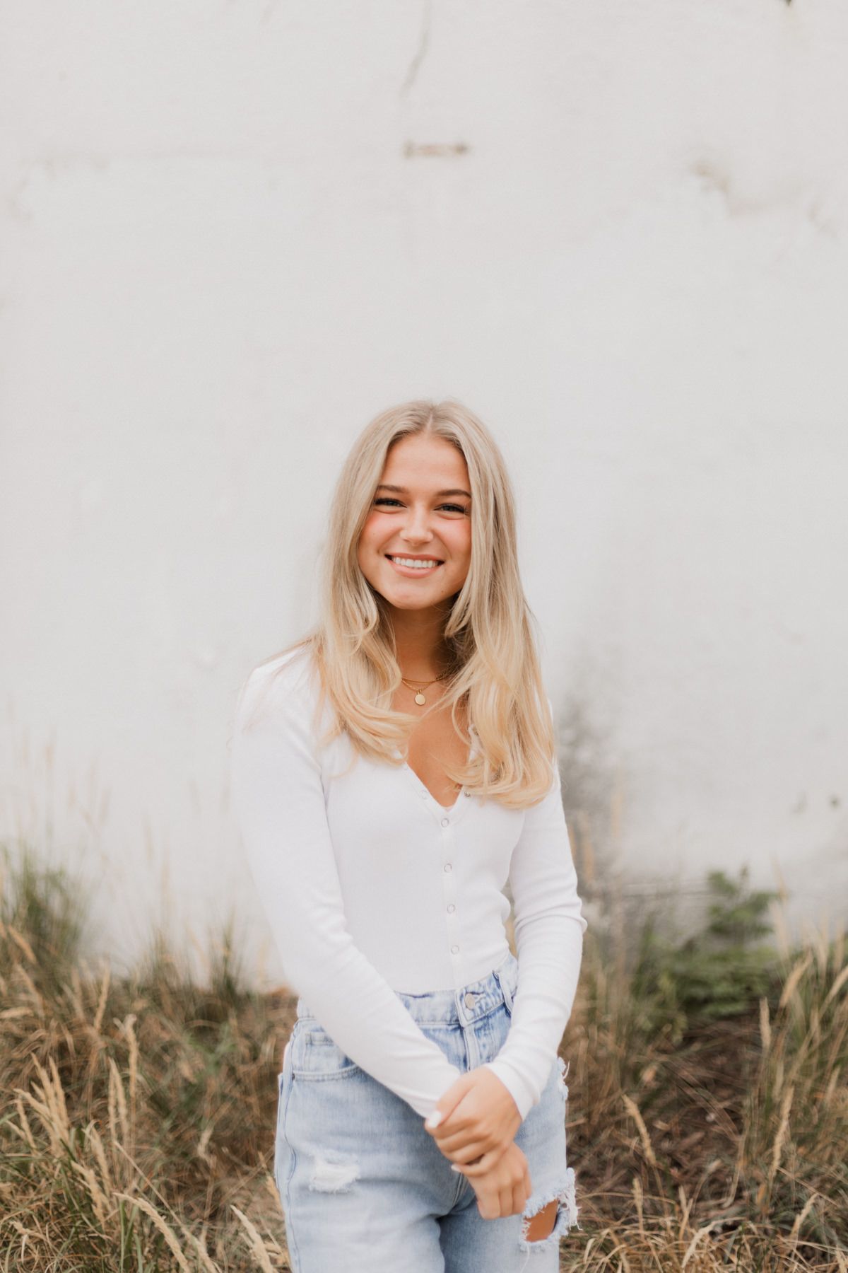 Blonde woman in white top and ripped jeans smiling against a white wall with dry grass.