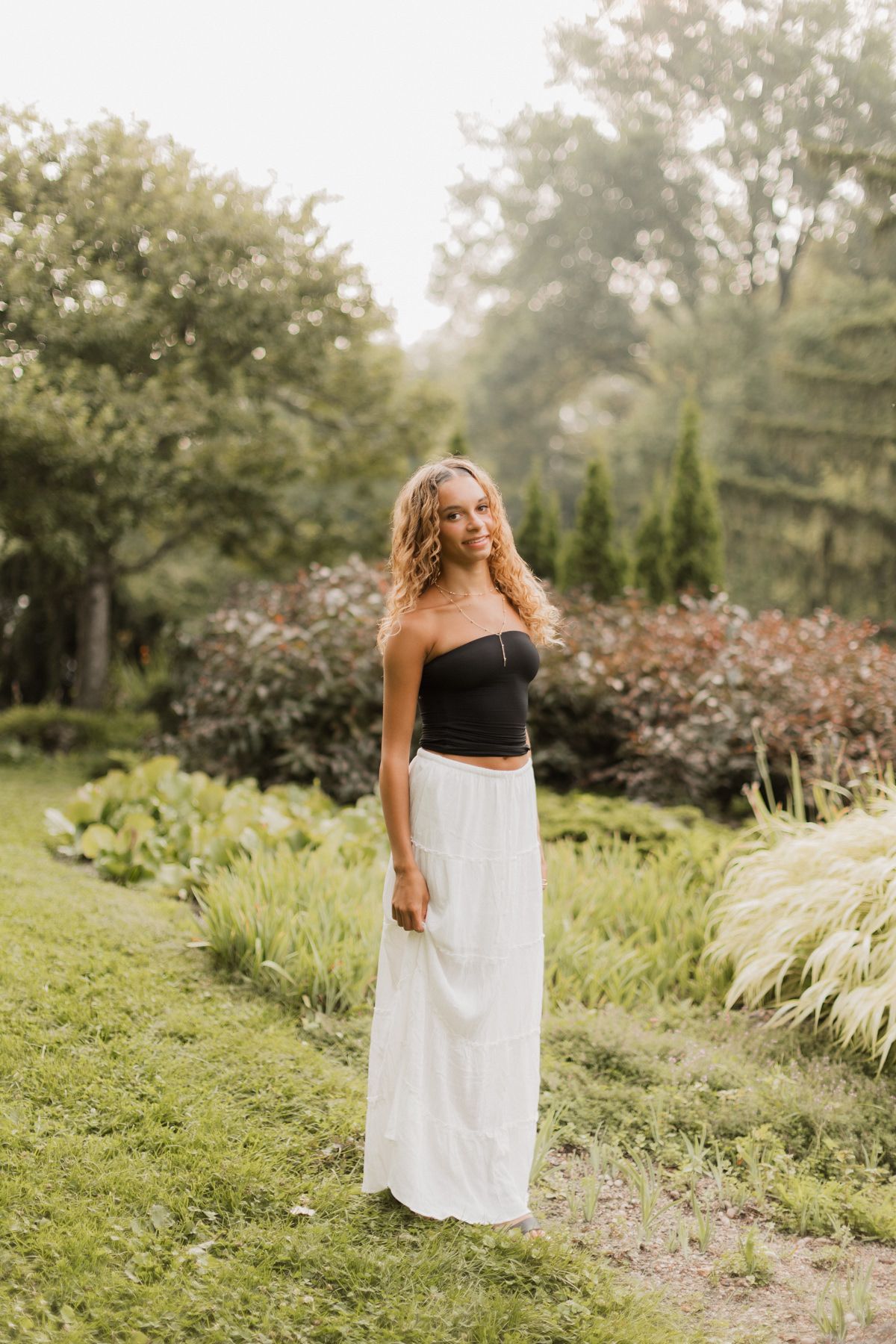 Young woman in black tube top and white maxi skirt posing in a lush green garden setting.