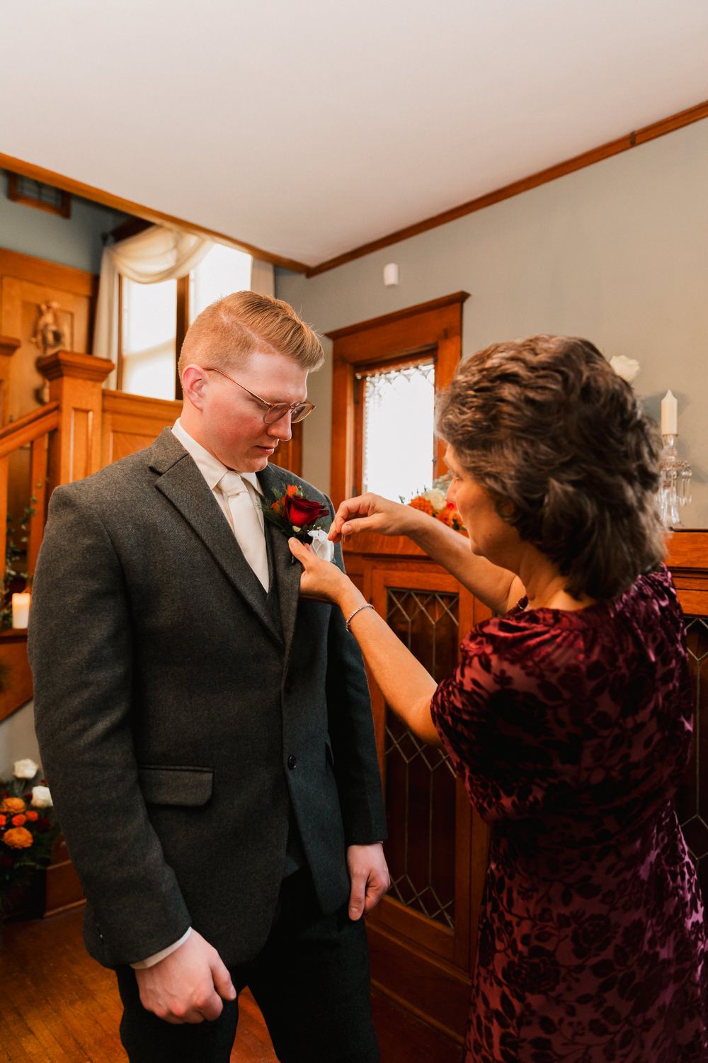 A boutonniere is pinned onto a dark suit jacket during wedding preparations in a warm-toned room.