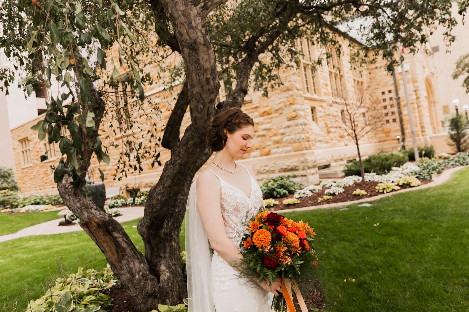 Wedding couple poses in front of historic stone building with trees and landscaped lawn.