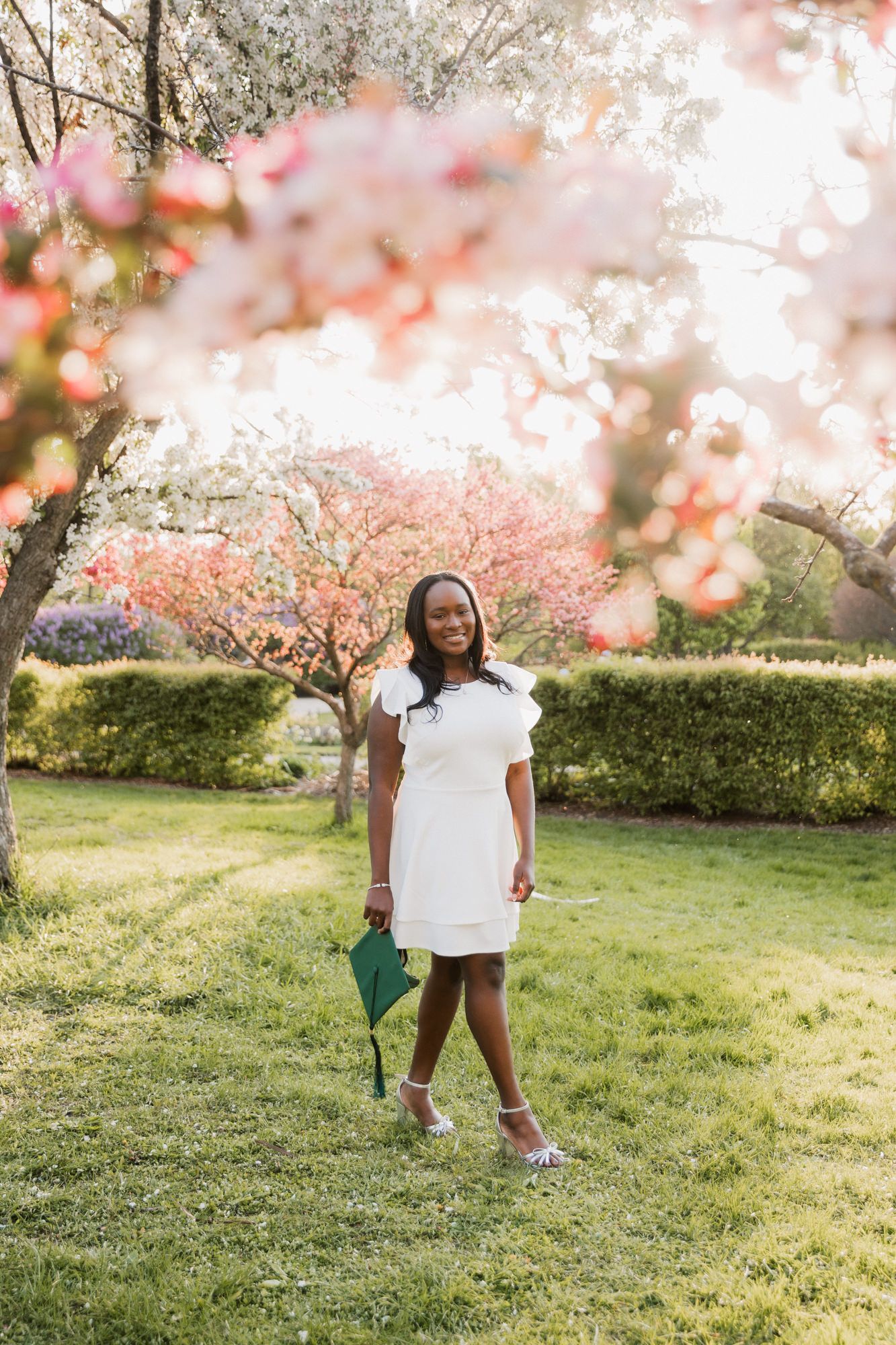 Young woman in white dress holding graduation cap poses among blooming cherry trees in a sunny garden.
