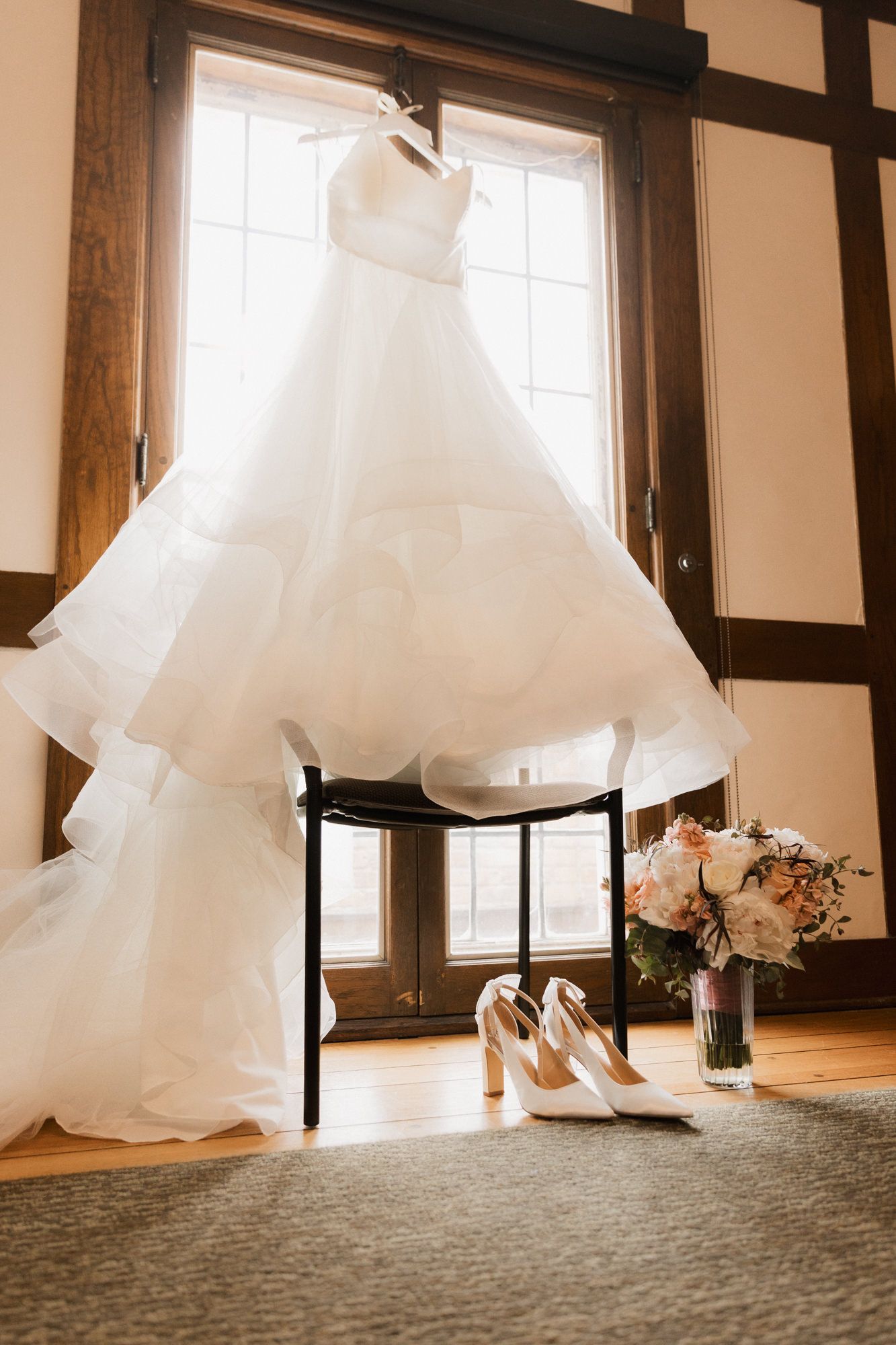 Elegant white ball gown wedding dress hanging by window with bridal heels and floral bouquet on wooden floor.