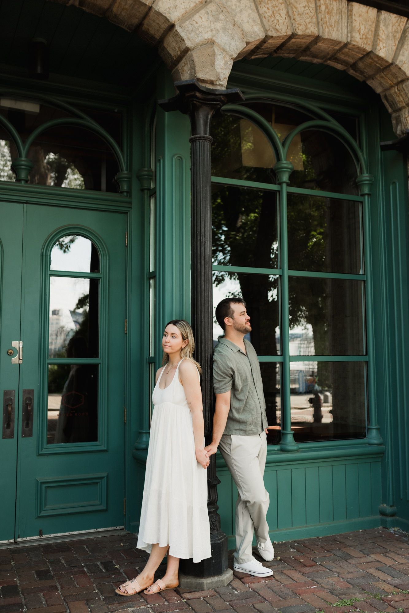 Couple holding hands standing back-to-back by green vintage doors on brick pavement in elegant outdoor setting.