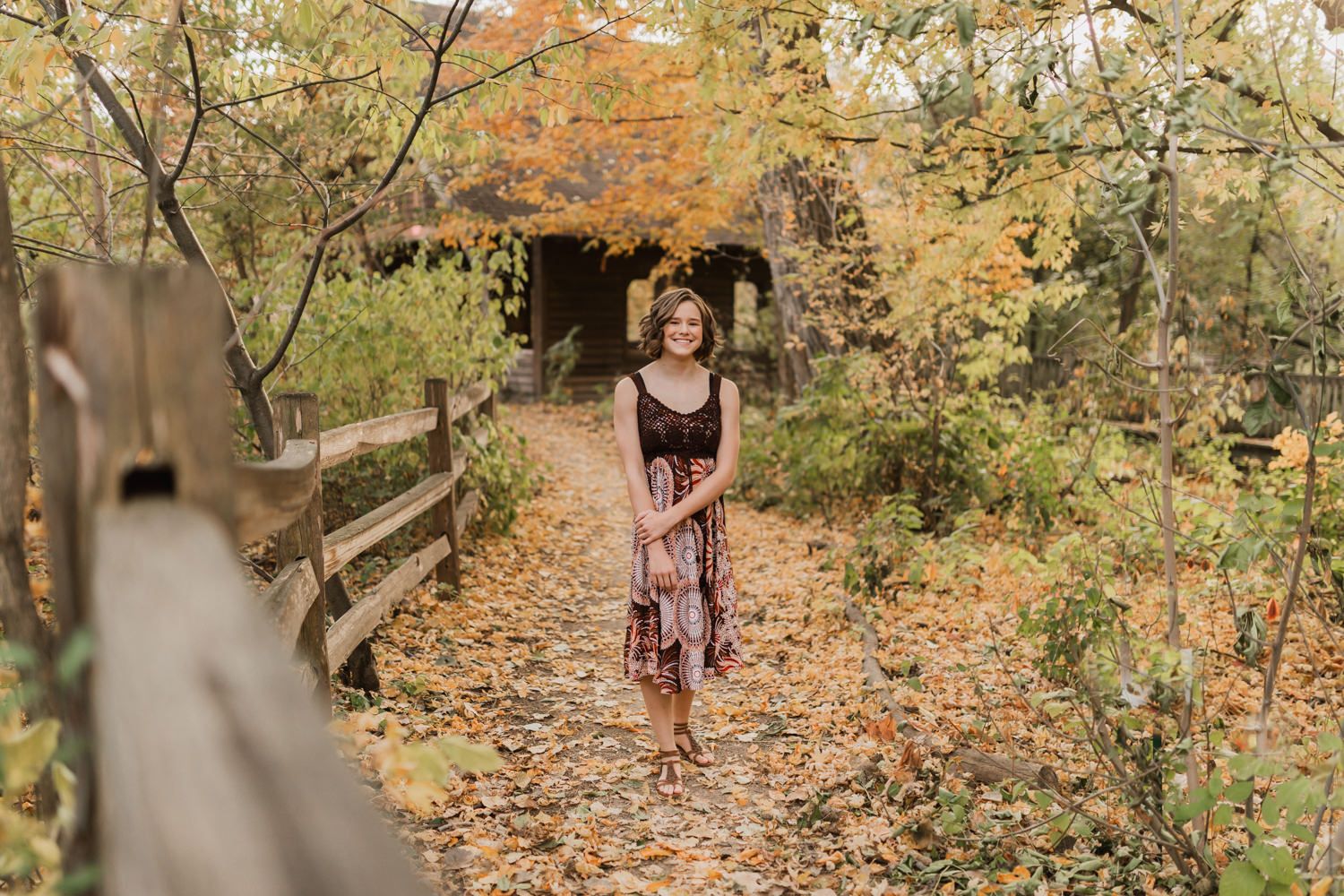 A person walks through a wooded path covered in fallen autumn leaves while trees display colorful fall foliage.