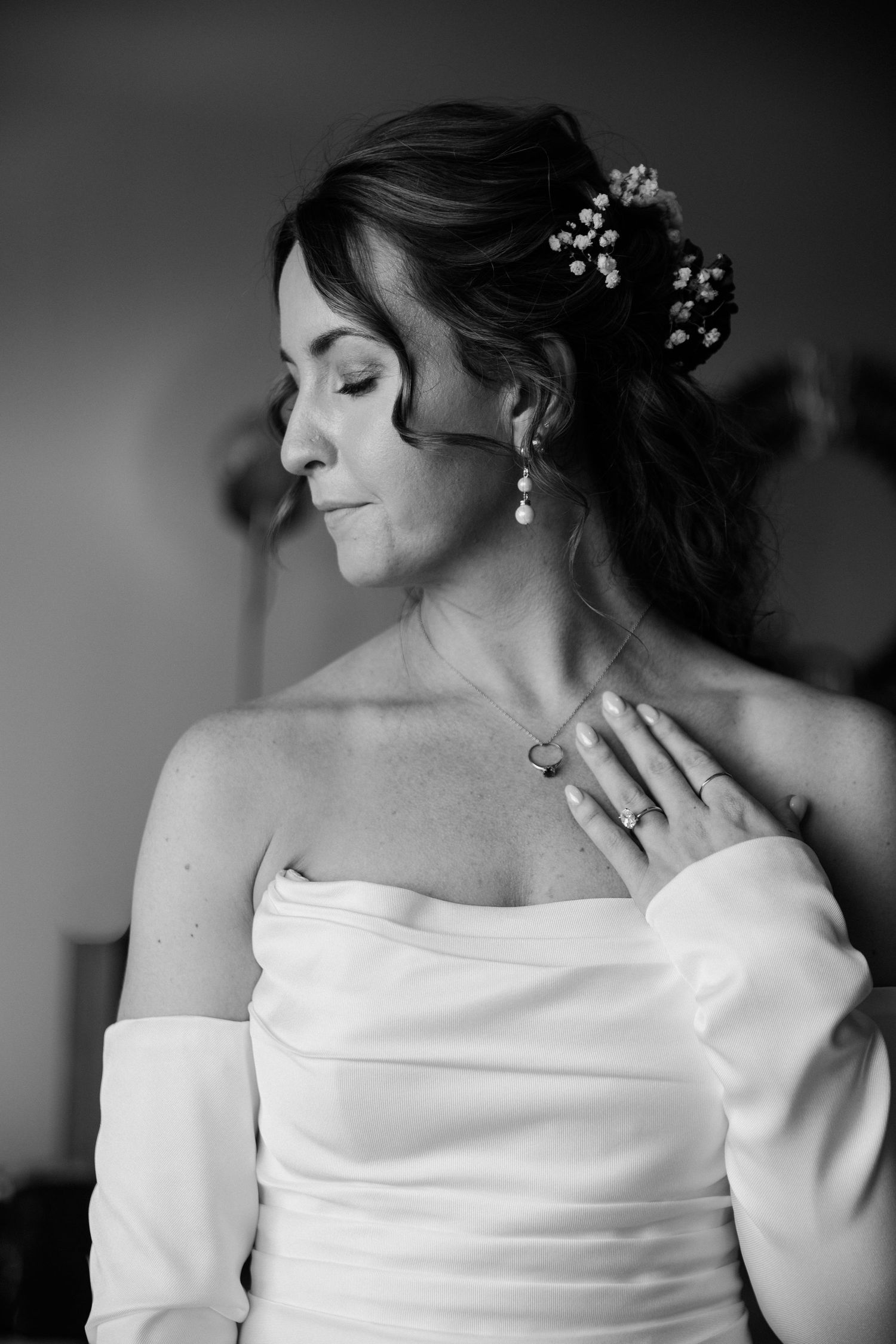 Black and white portrait of a bride in an off-the-shoulder satin gown with pearl earrings and baby's breath in her hair.