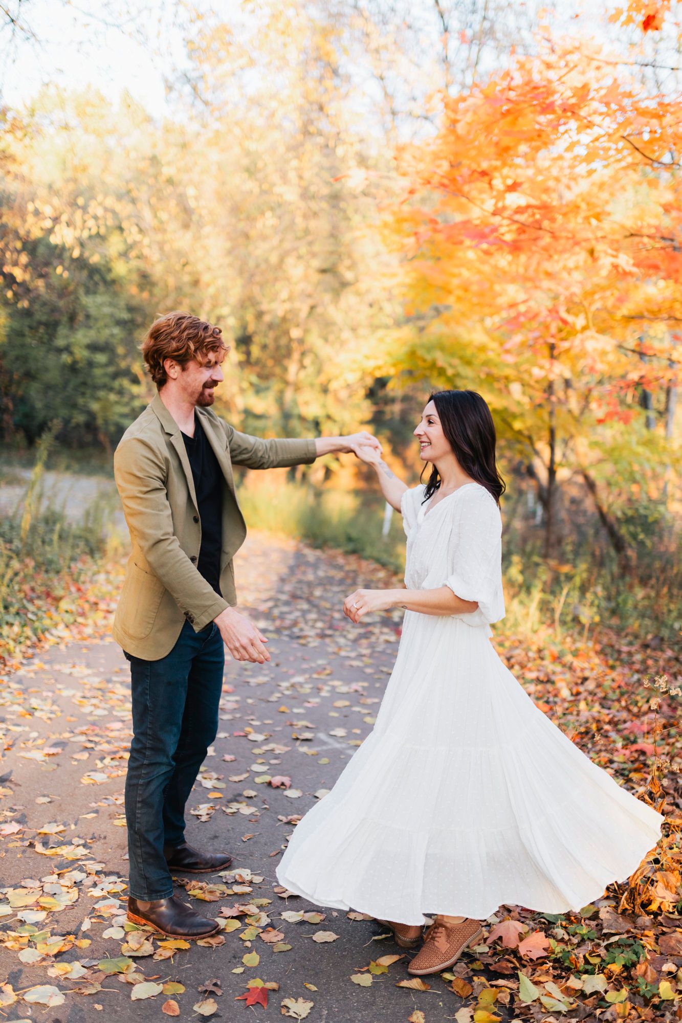 A couple dances playfully on an autumn path covered with fallen orange leaves.