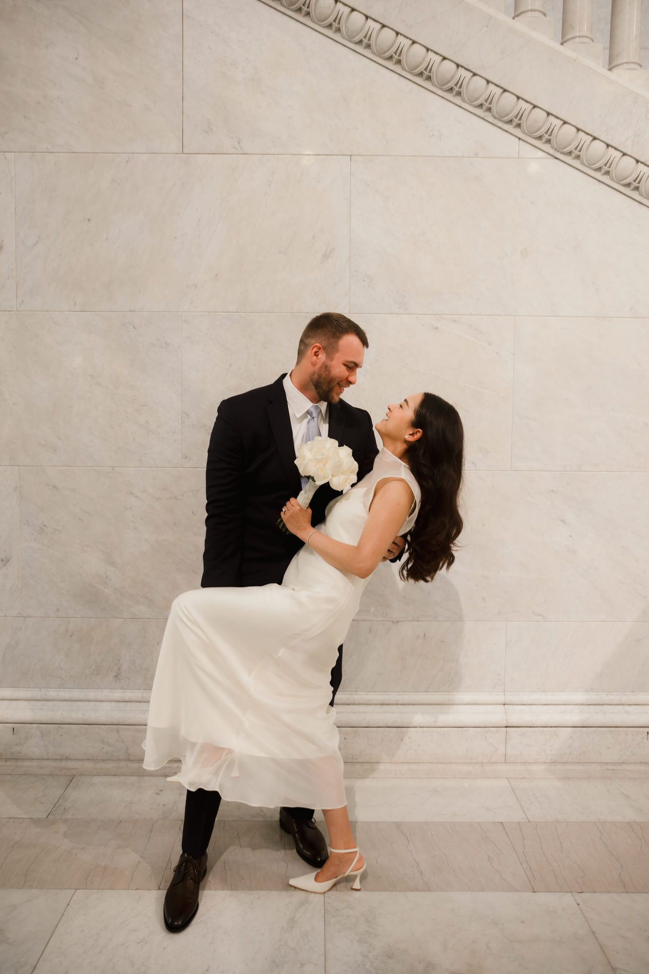 A groom dips his bride in a white dress and heels against a marble wall, sharing a joyful romantic moment.