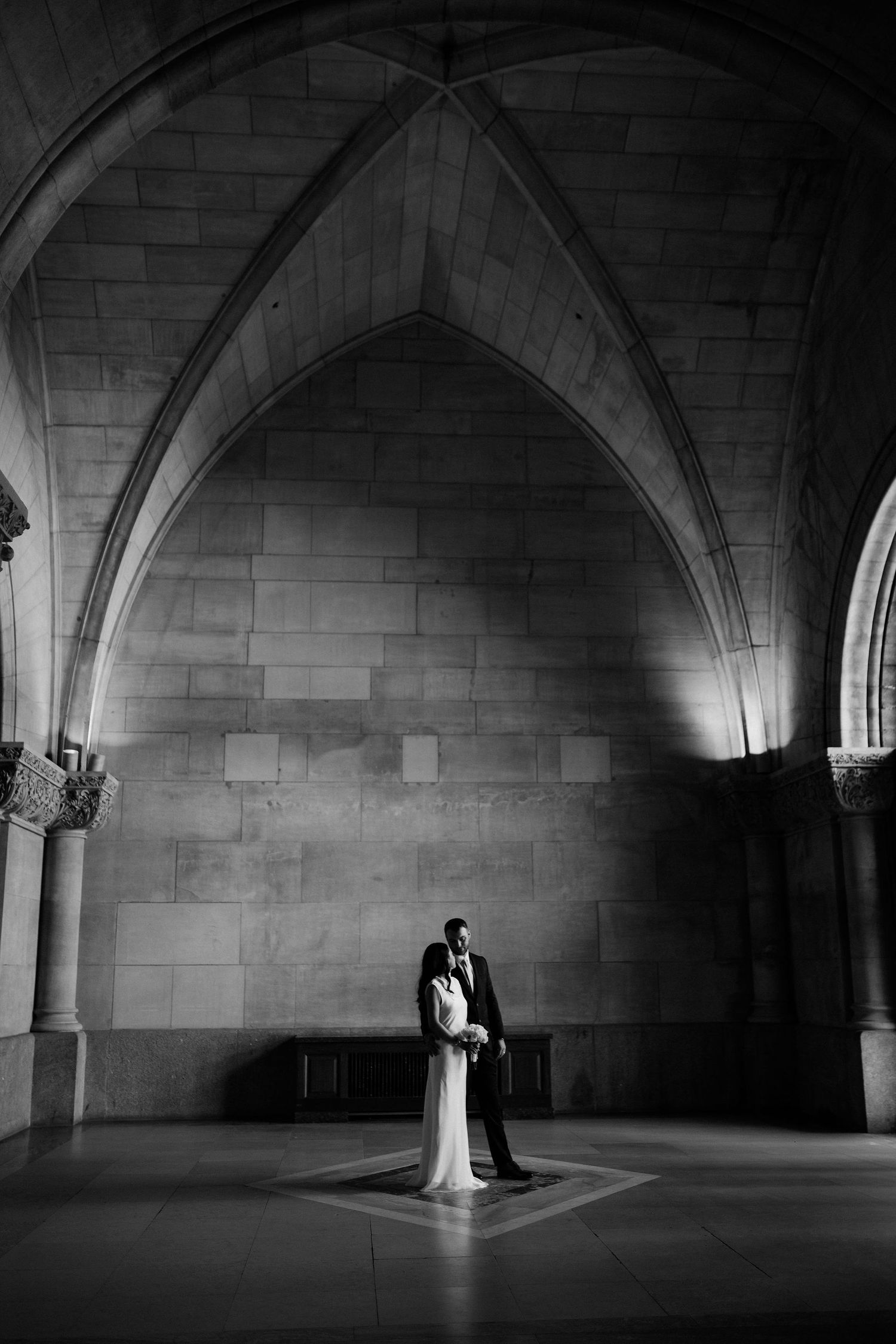 Black and white wedding photo of bride and groom embracing under Gothic stone arches in an elegant cathedral.