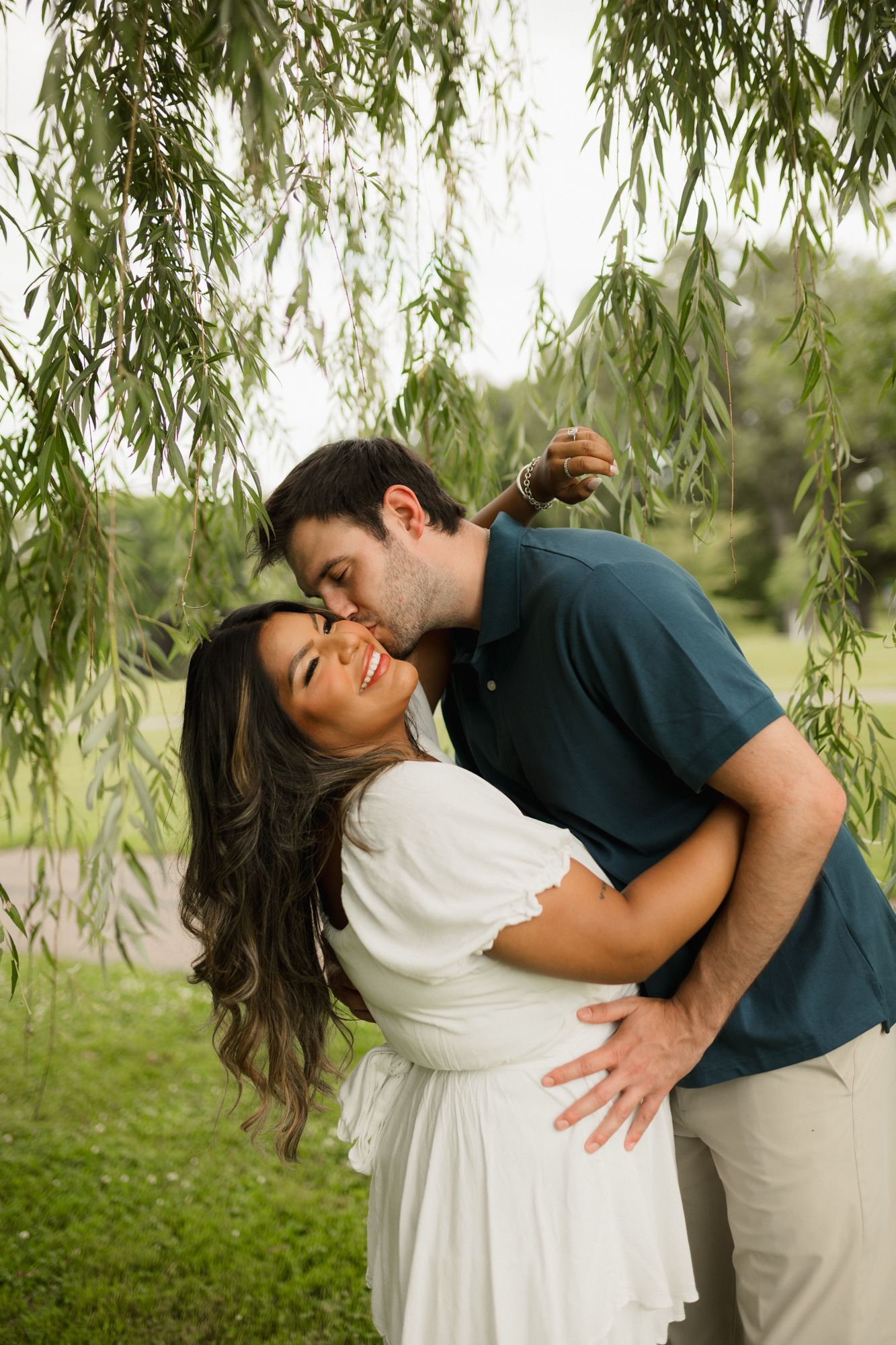 A couple shares a romantic kiss under a weeping willow tree, with the woman in a white dress smiling joyfully.