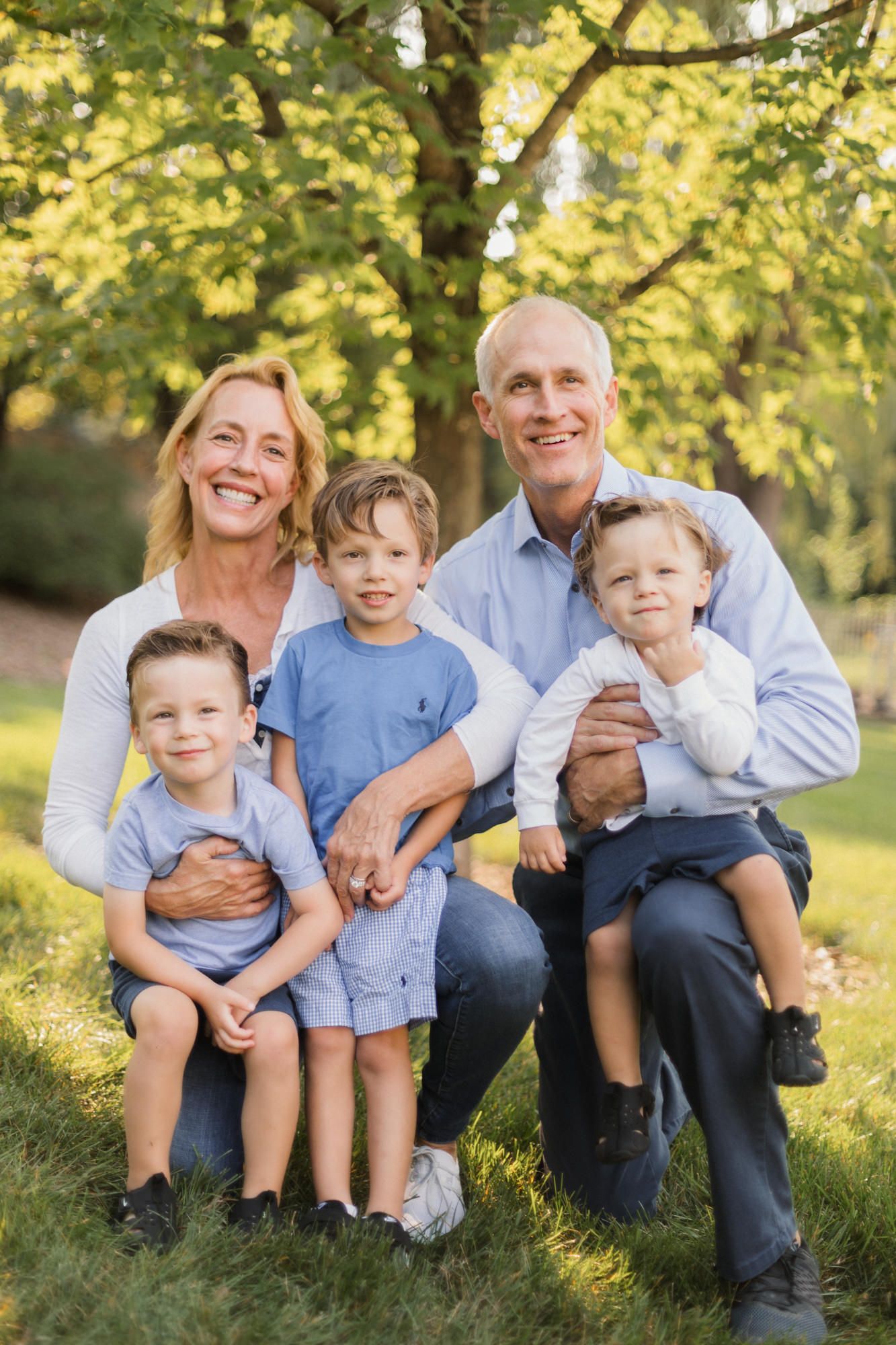 Family portrait of two parents with three young boys in blue outfits, smiling outdoors under green trees.