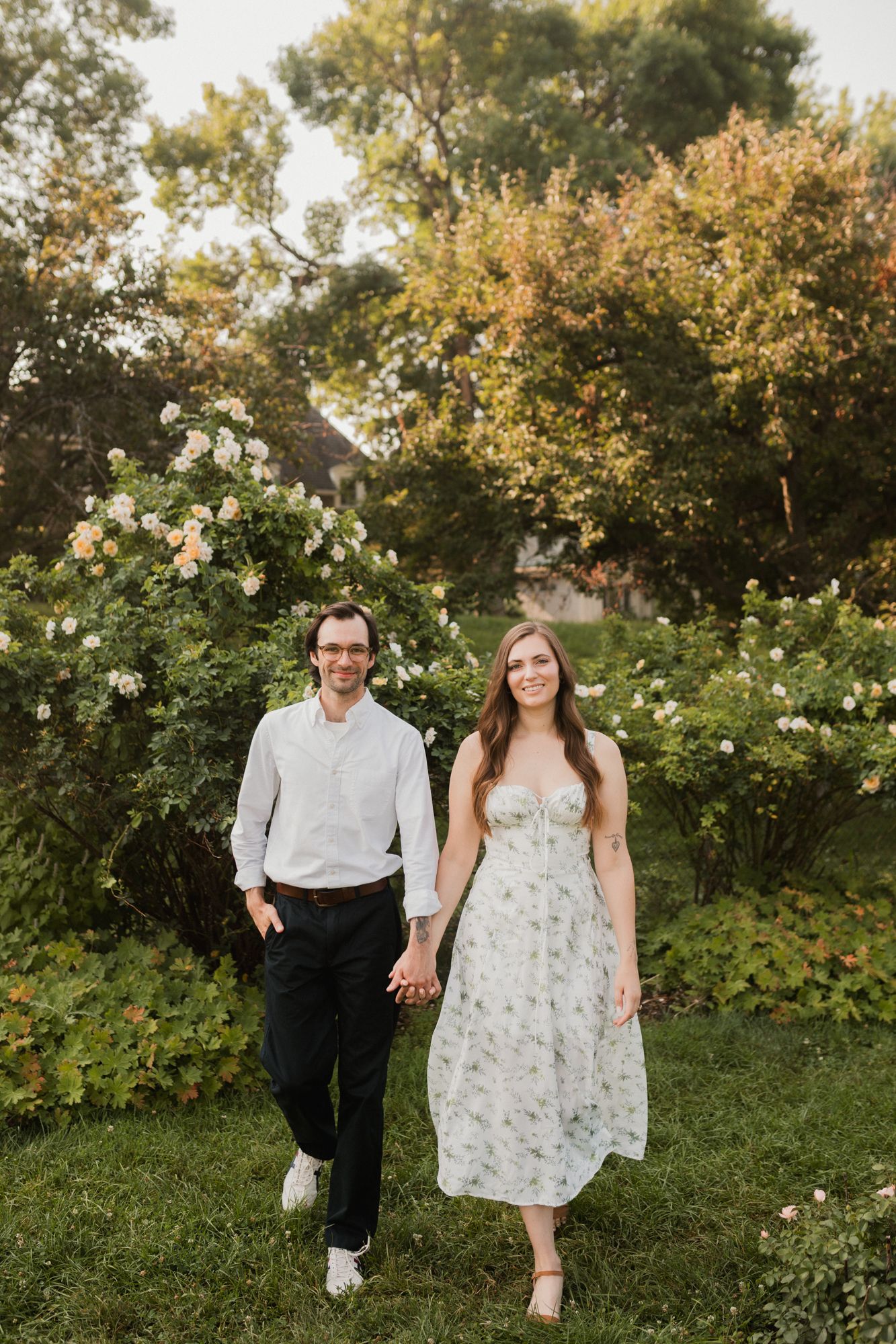 A couple holds hands walking through a lush garden, woman in floral dress, man in white shirt and black pants.