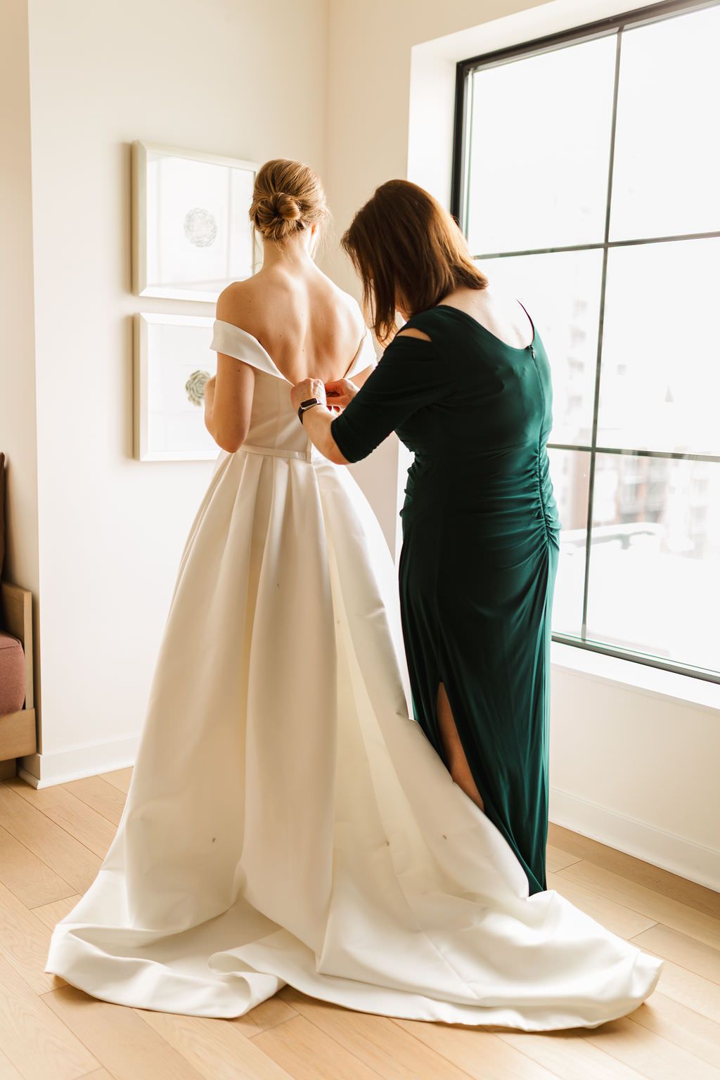 Helper assists bride with wedding dress details near large window in preparation room.