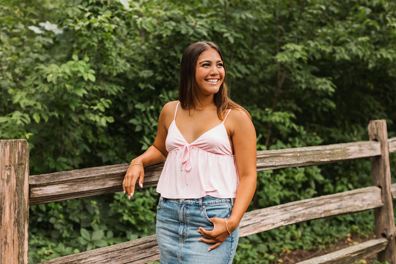 A person in a white tank top and denim shorts poses outdoors near a wooden fence amid lush greenery.