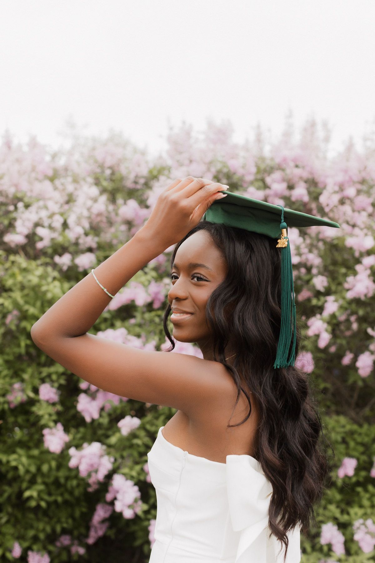 Young Black woman in white dress adjusts green graduation cap, smiling outdoors near blooming pink flowers.