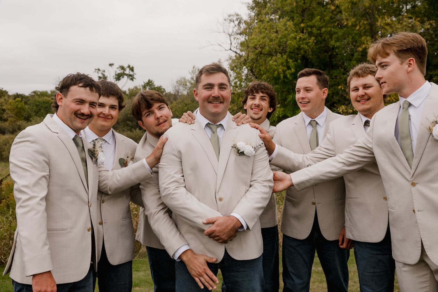 Groom in beige suit smiles as groomsmen in matching suits playfully pose together outdoors at a wedding.