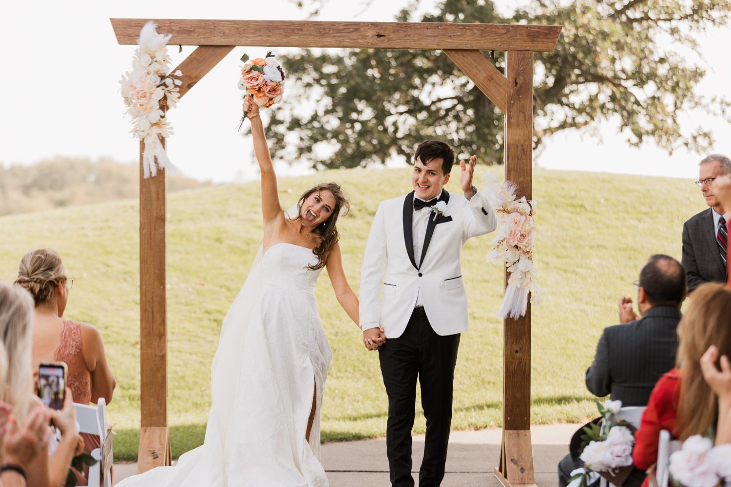 A joyful celebration under a rustic wooden wedding arch decorated with white flowers in an outdoor countryside setting.