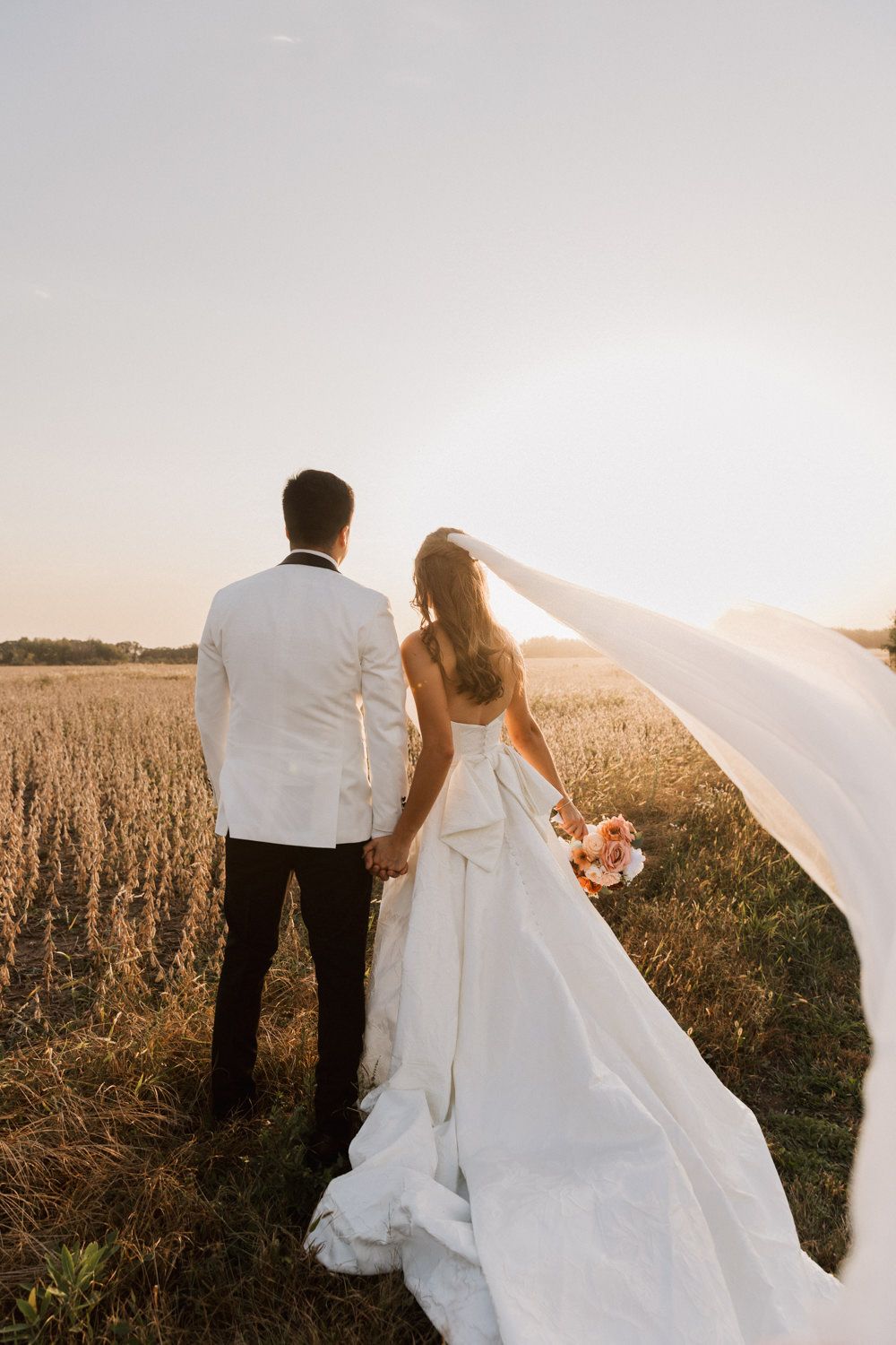 A romantic sunset wedding photo of a couple in a field with the bride's veil flowing in the wind.