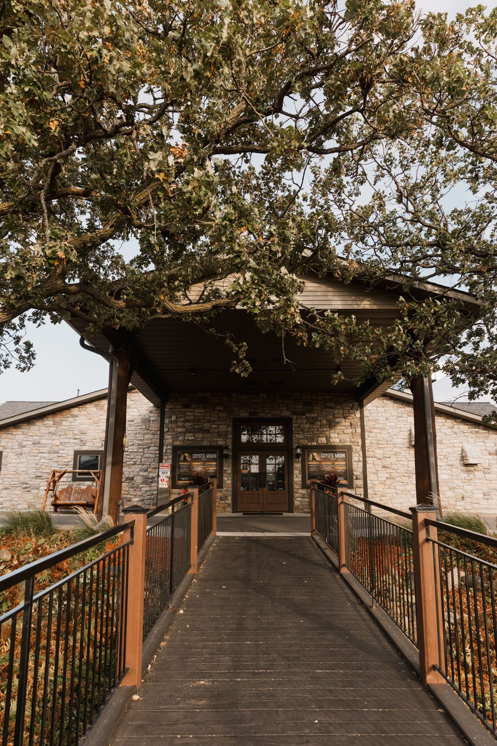 A wooden ramp walkway with rustic railings leads up to a stone building under the shade of large oak trees.