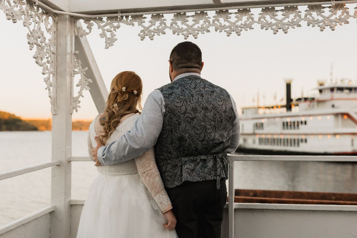 Couple looks out over river at sunset from decorated wedding venue deck with riverboat in background.