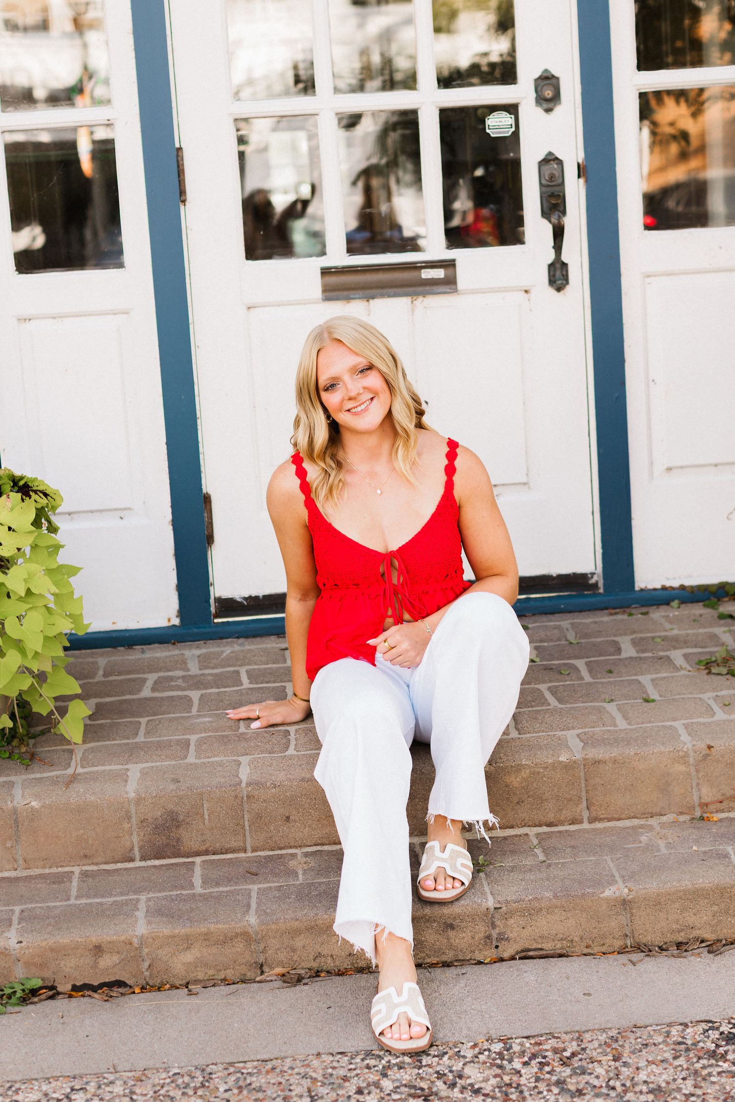 Woman in red tank top and white pants sits casually on white porch steps during outdoor photo session.