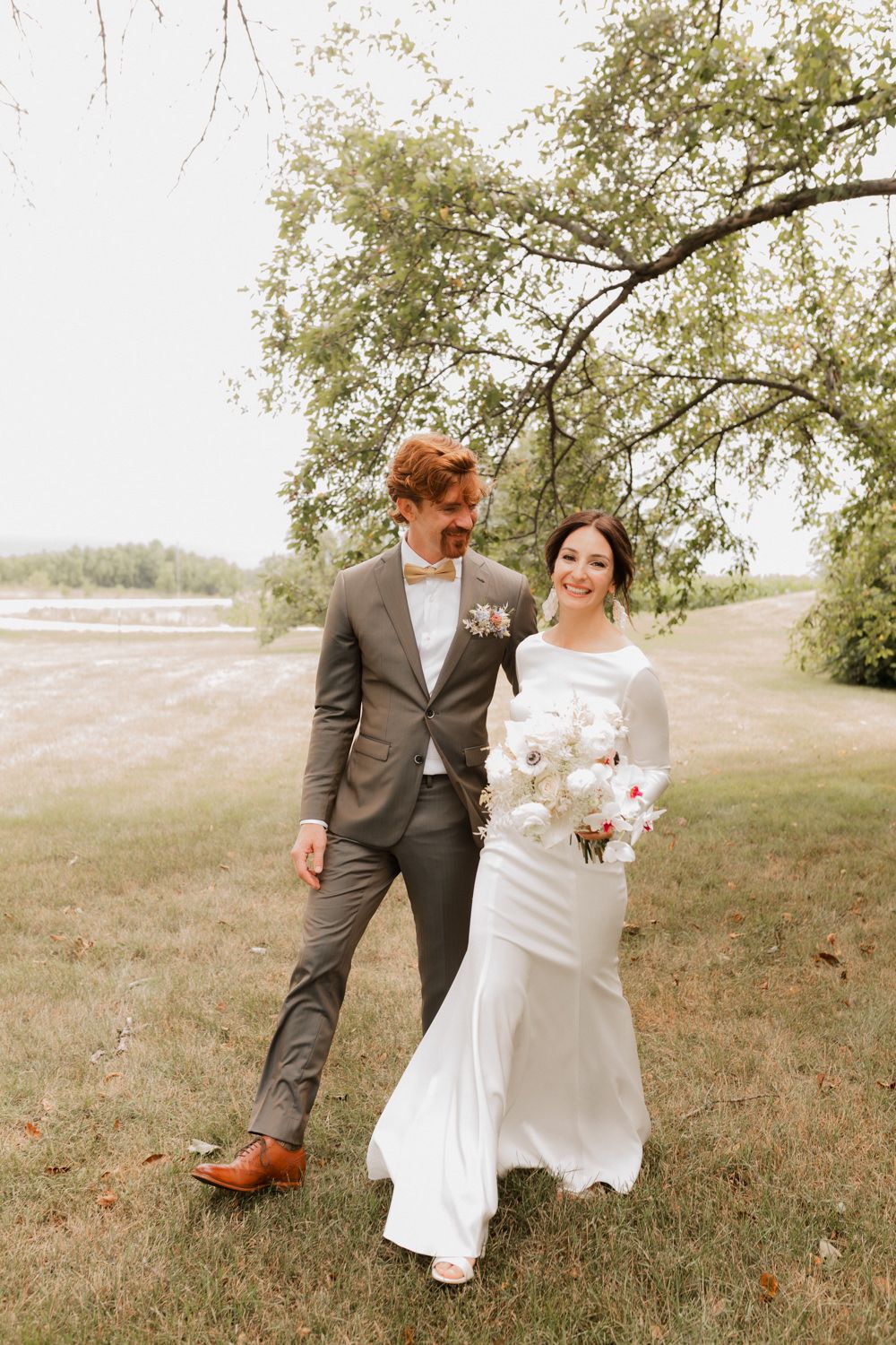 A couple in wedding attire stands together on grassy terrain under a tree branch, with the bride holding a bouquet.
