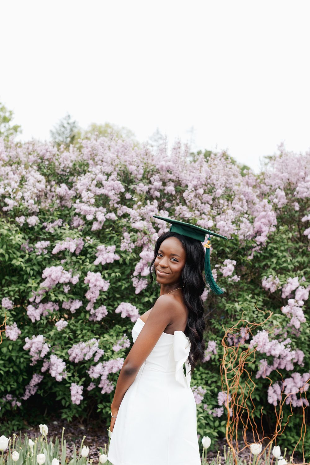 Graduate in white dress poses gracefully against backdrop of pink flowering trees.