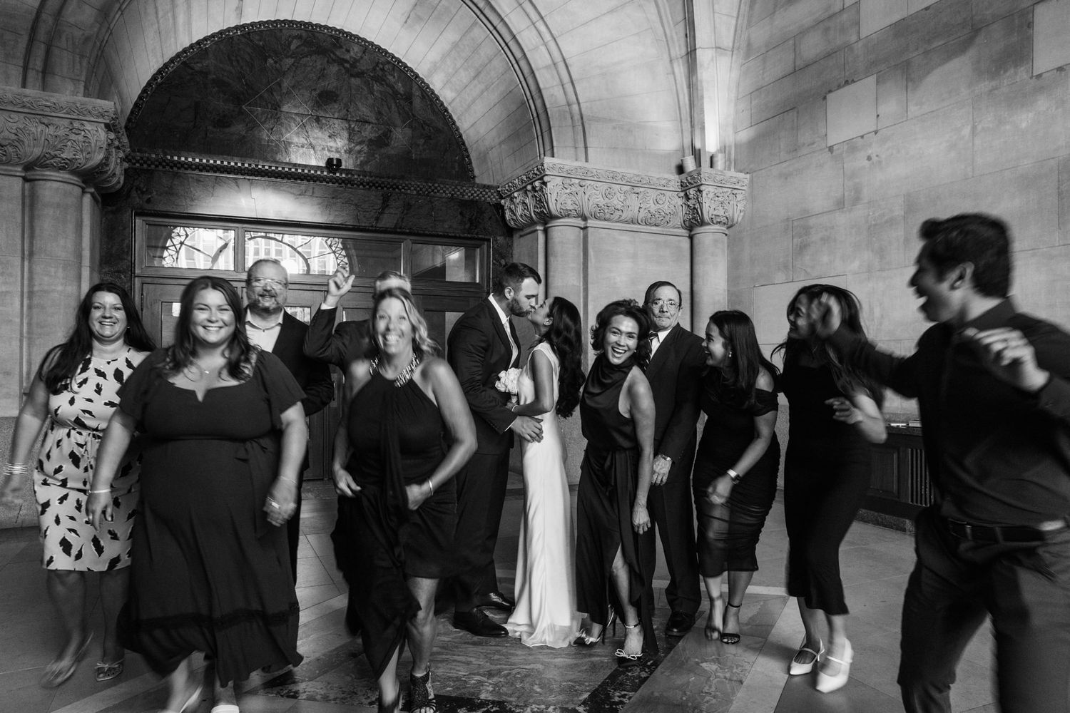 Black and white wedding photo of bride, groom, and guests posing under ornate stone archway.